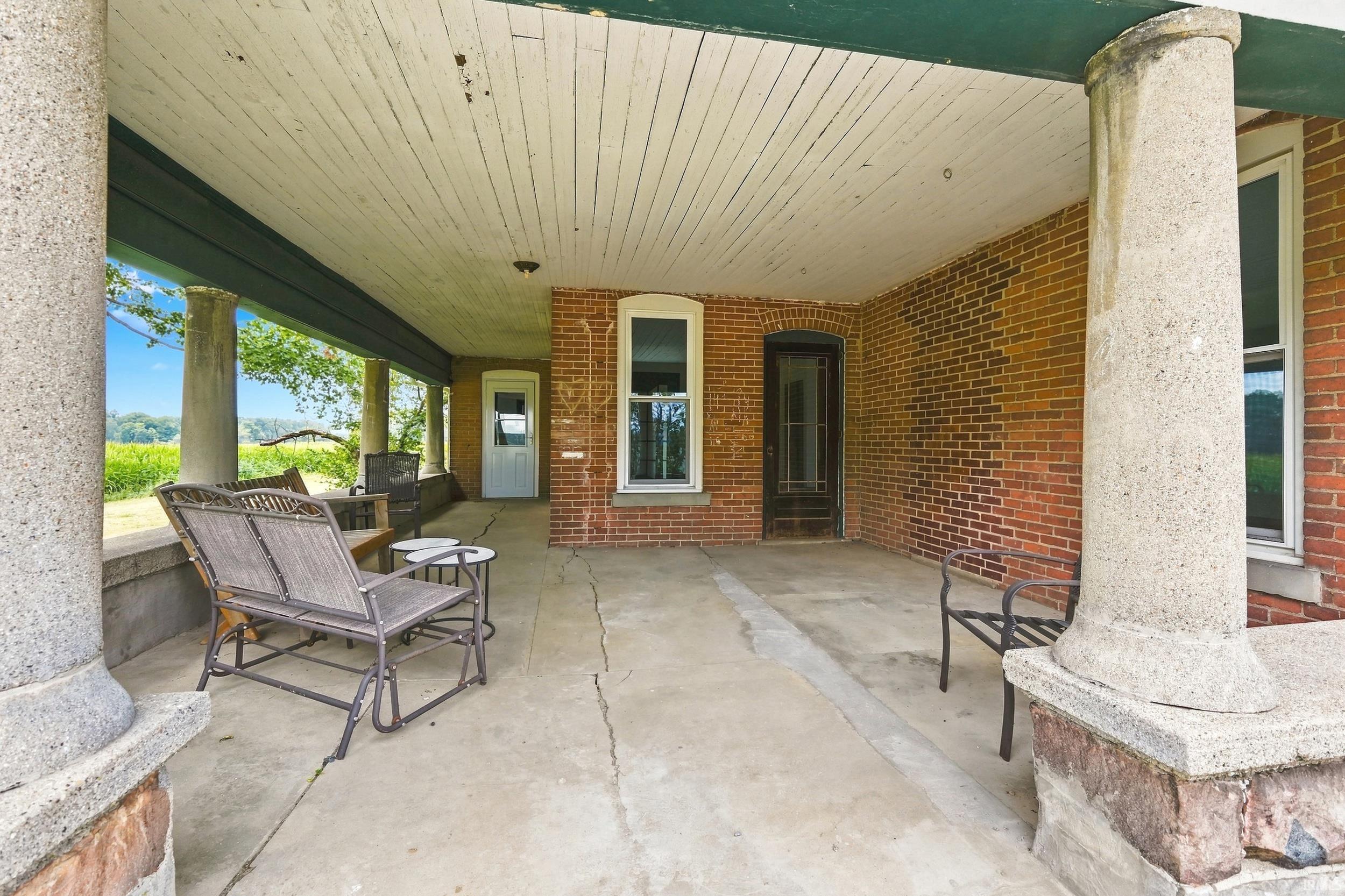 Covered front porch featuring concrete flooring, stone support columns, brick exterior siding, and a wooden plank ceiling