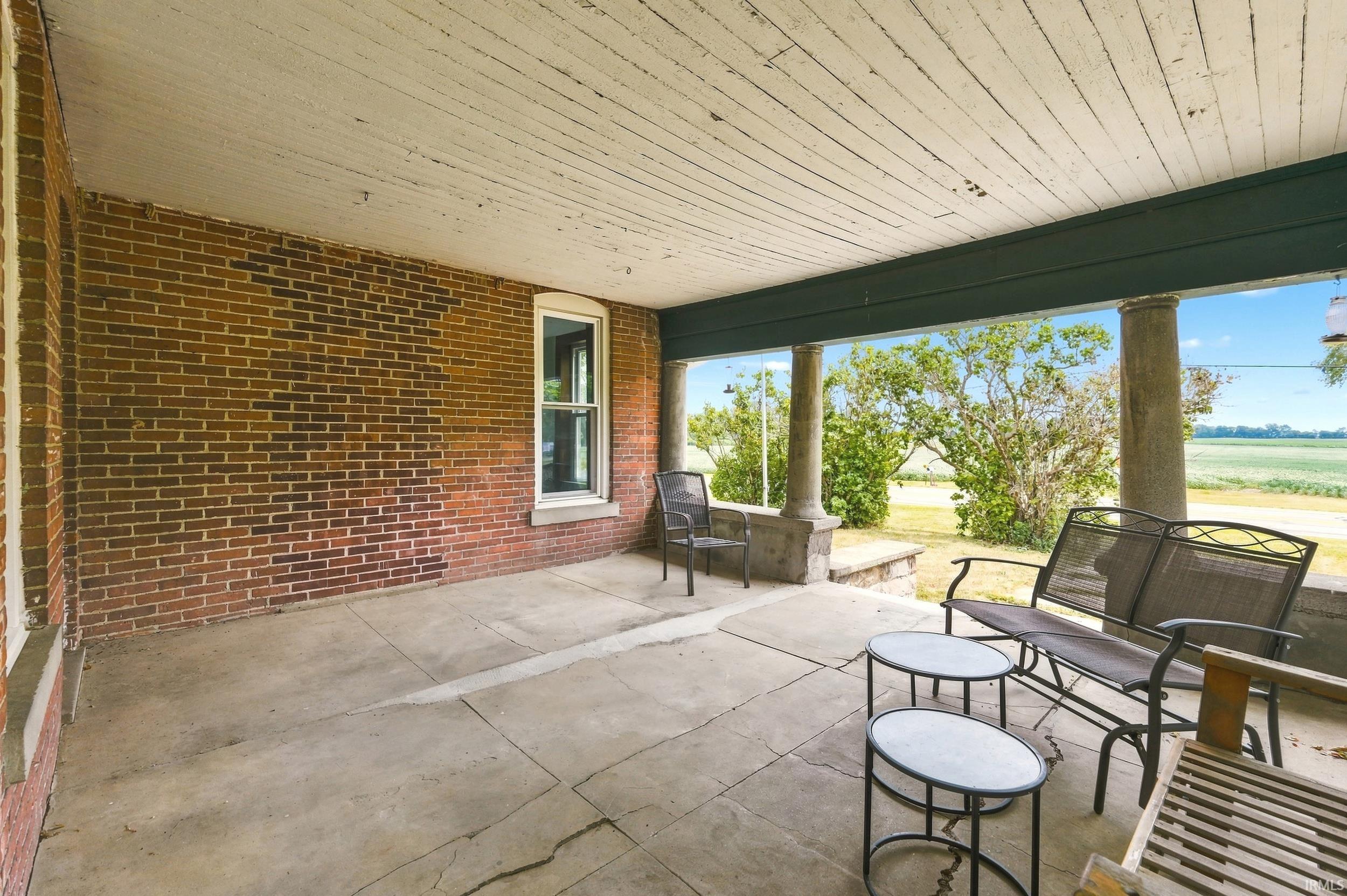 Covered porch featuring a brick exterior wall, a white-framed window, concrete flooring, and substantial support columns