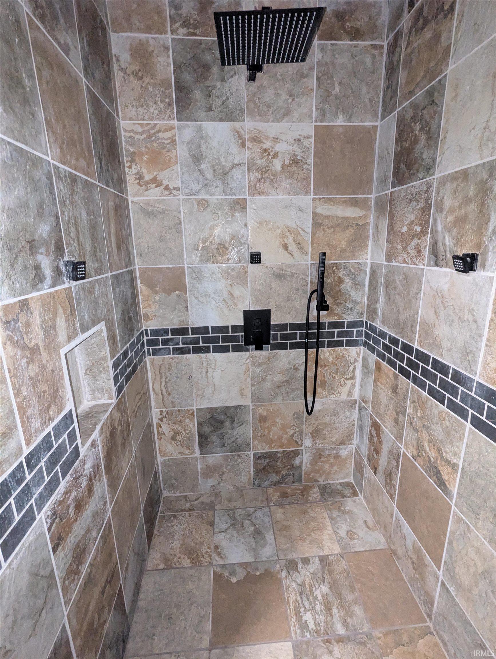 Tiled shower stall featuring a matte black rainfall showerhead, handheld shower fixture, and control panel