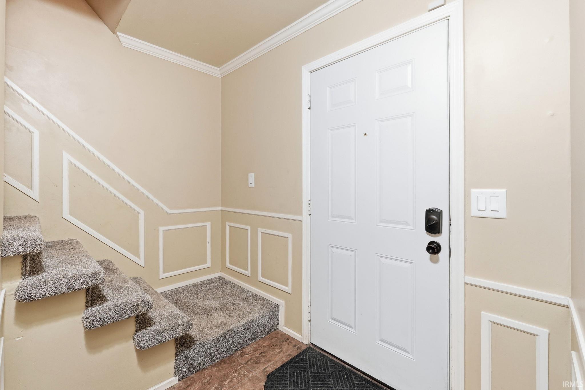 Entryway featuring a white paneled door with black hardware, wainscoting detailing, crown molding, and carpeted stairs