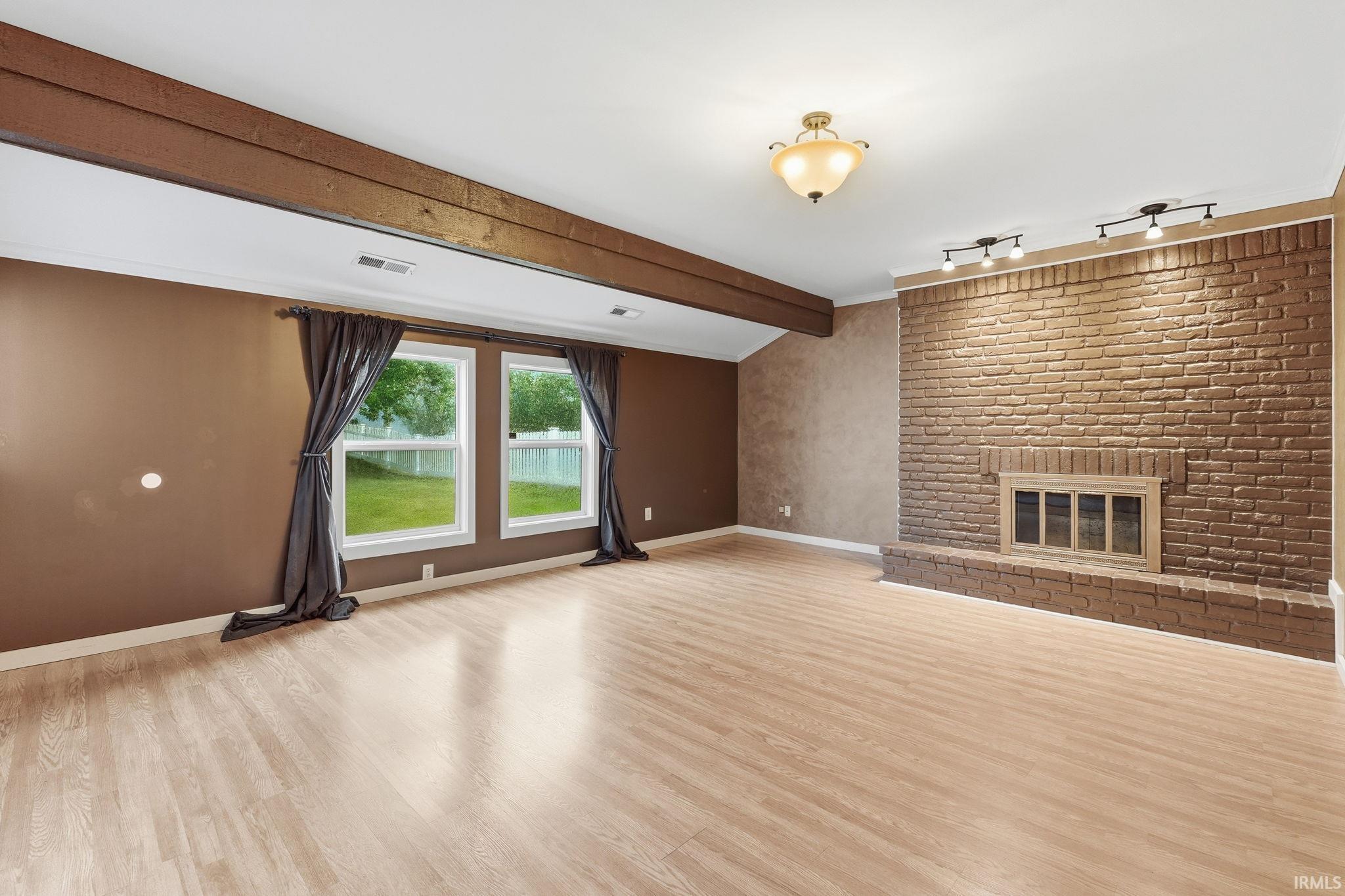 Wood-finish flooring throughout, prominent brick-finish fireplace, two large windows, wood beam ceiling accent, and ceiling-mounted track lighting
