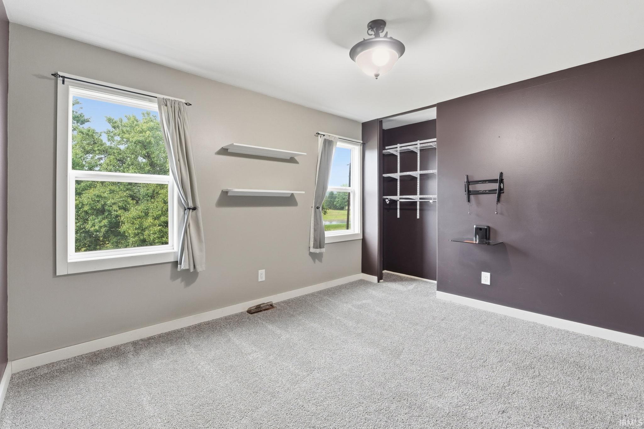 Plush gray carpeting throughout, featuring two windows with white trim, built-in closet shelving, and a wall-mounted light fixture