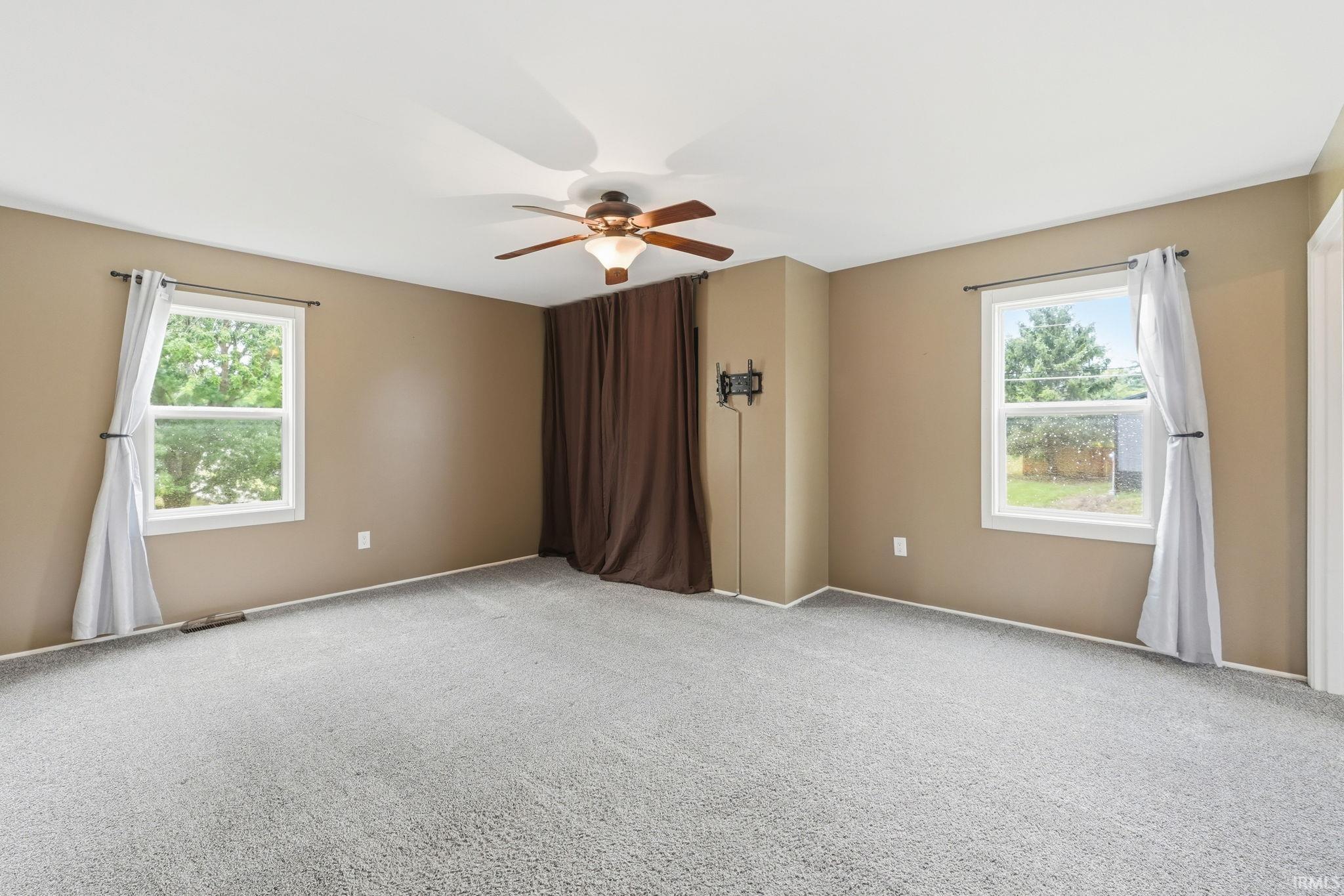 Spacious room featuring light gray carpeting, neutral-toned walls, two windows, and a ceiling fan with integrated lighting
