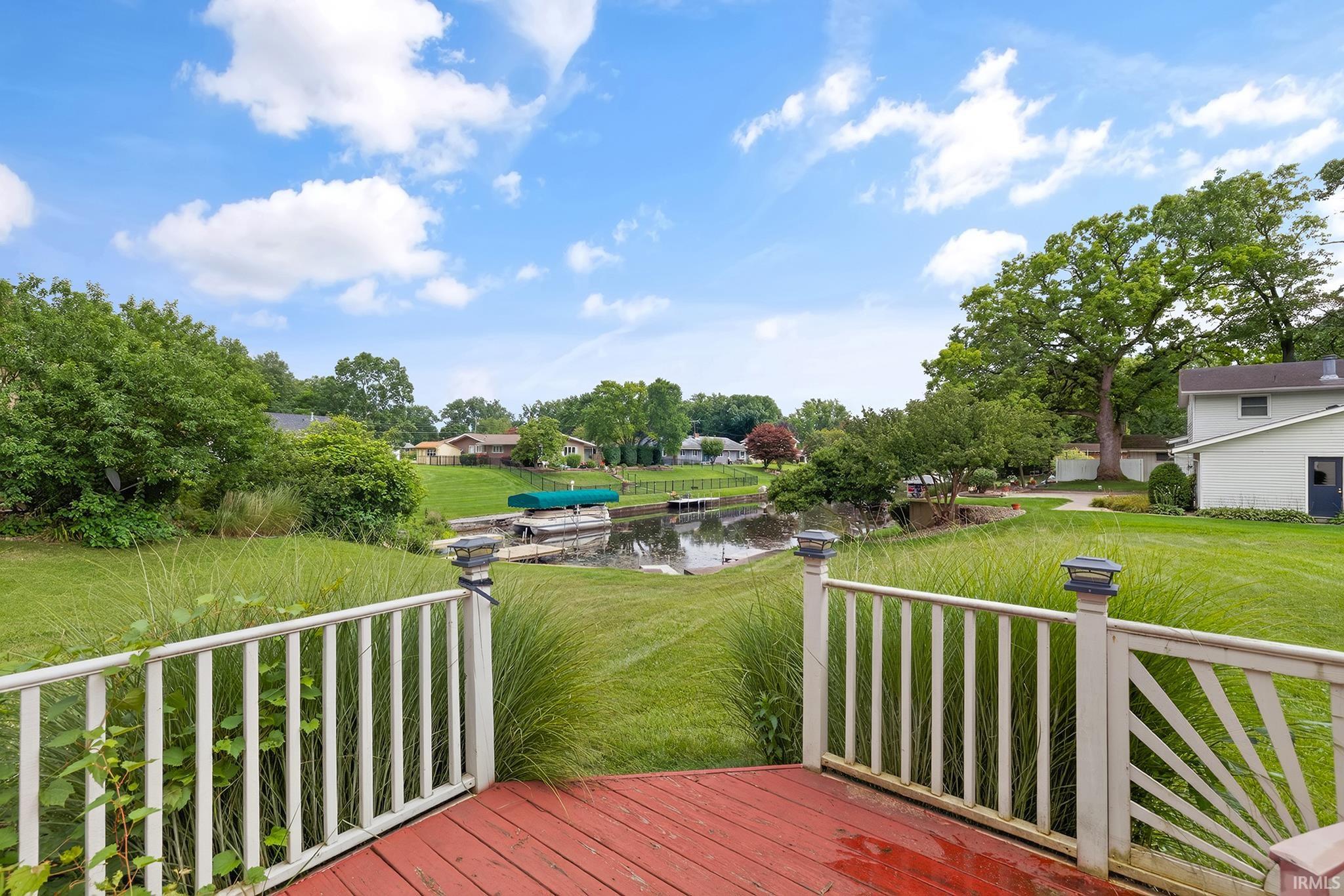 Red-stained deck with white railing featuring solar post caps, overlooking a waterway with a boat dock and boat lift, surrounded by a green lawn and mature trees