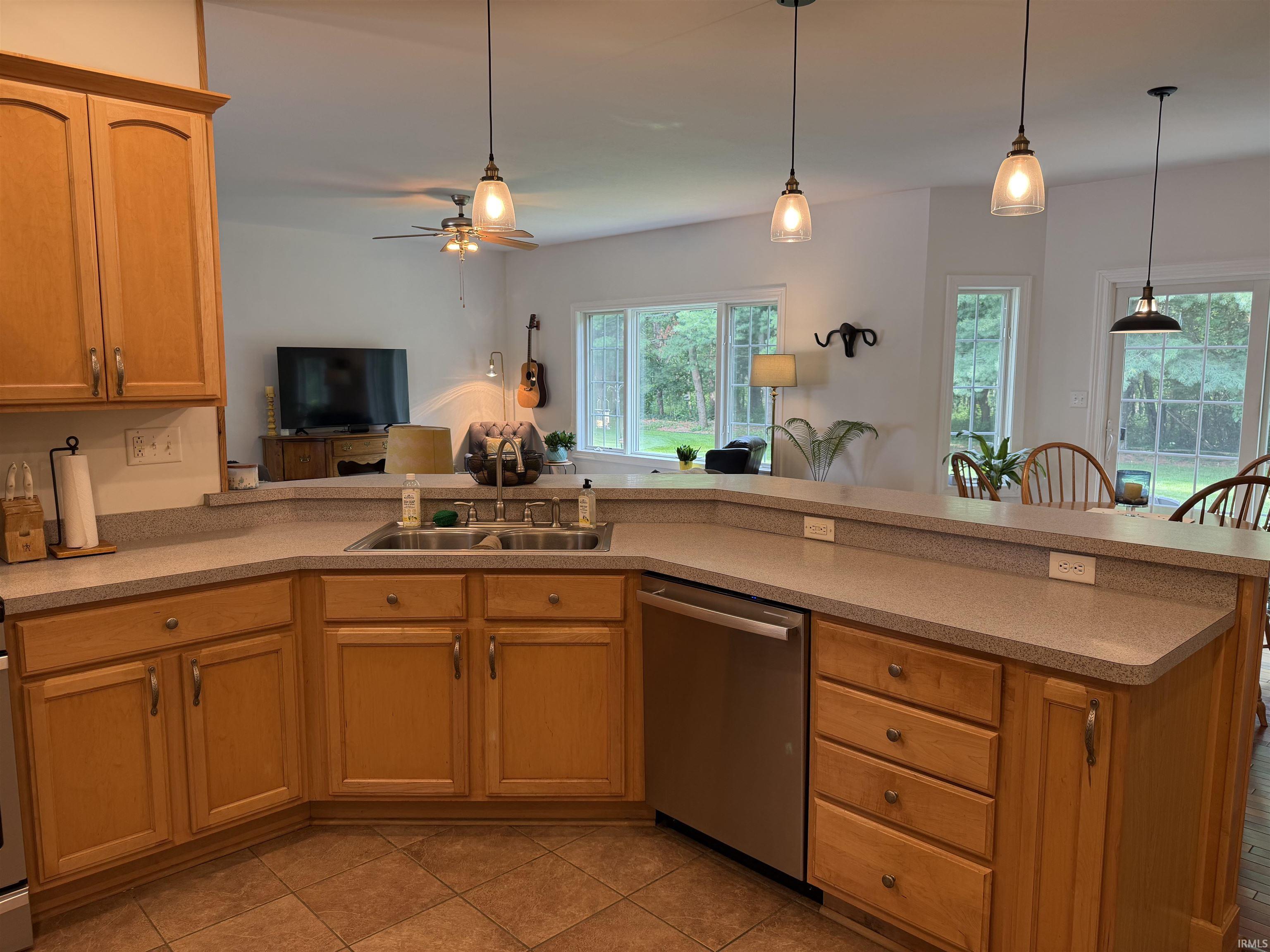 Open-concept kitchen featuring light-toned wood cabinetry, a double basin sink, stainless steel dishwasher, and a breakfast bar with pendant lighting