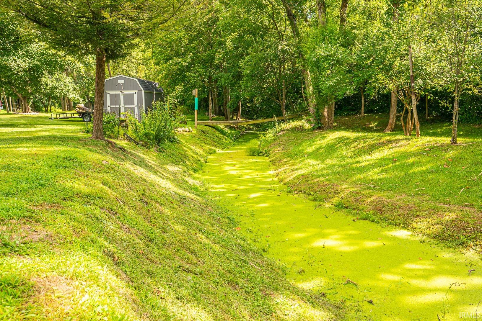 Lush green landscape featuring a channel of water, mature trees, a gray storage shed, and a wooden footbridge