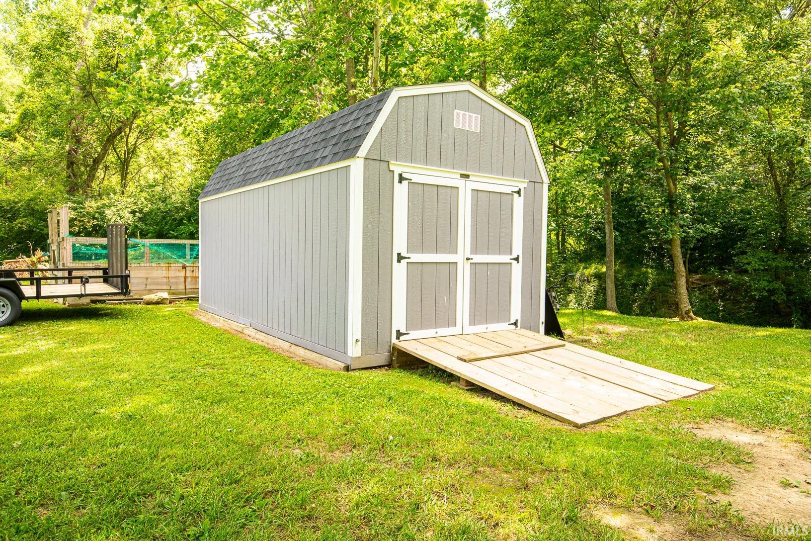 Detached barn-style shed featuring gray siding, white trim, a black shingle roof, and double doors with an integrated wooden ramp