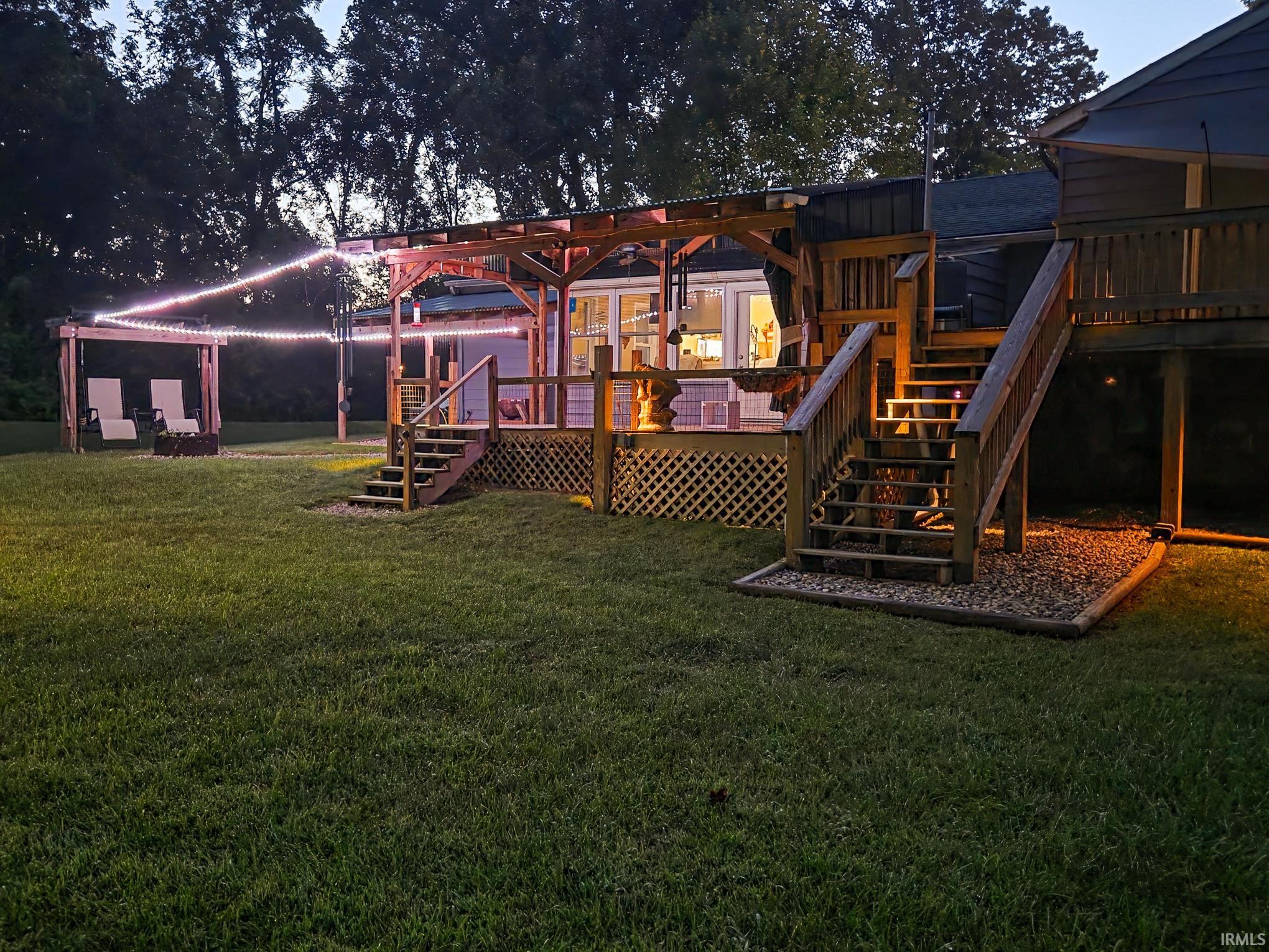 Rear exterior featuring a multi-level wooden deck with lattice skirting, wood pergola with string lights, and a spacious green lawn