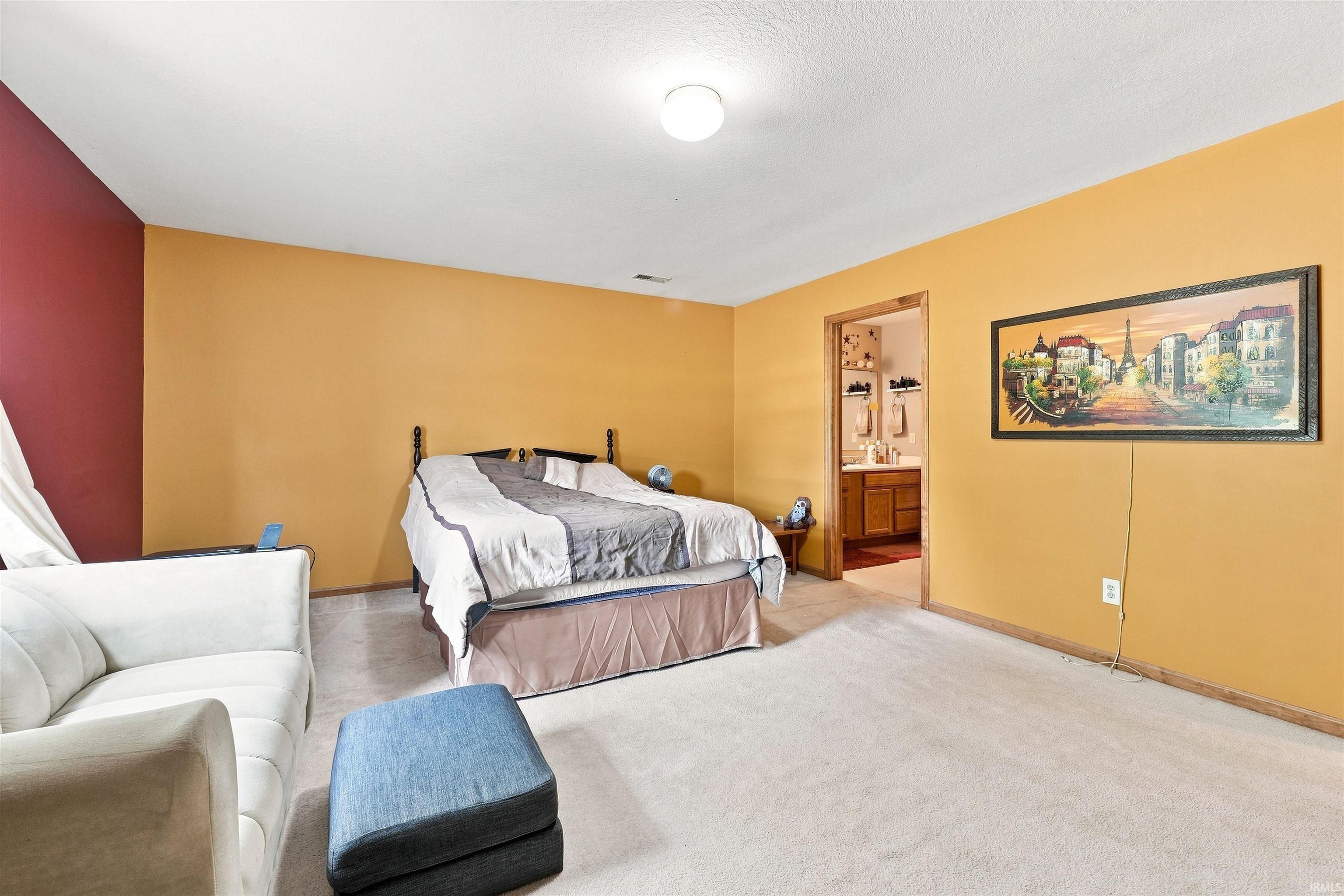 Spacious primary bedroom with warm golden-toned walls, carpeted flooring, and abundant natural light from recessed ceiling fixtures