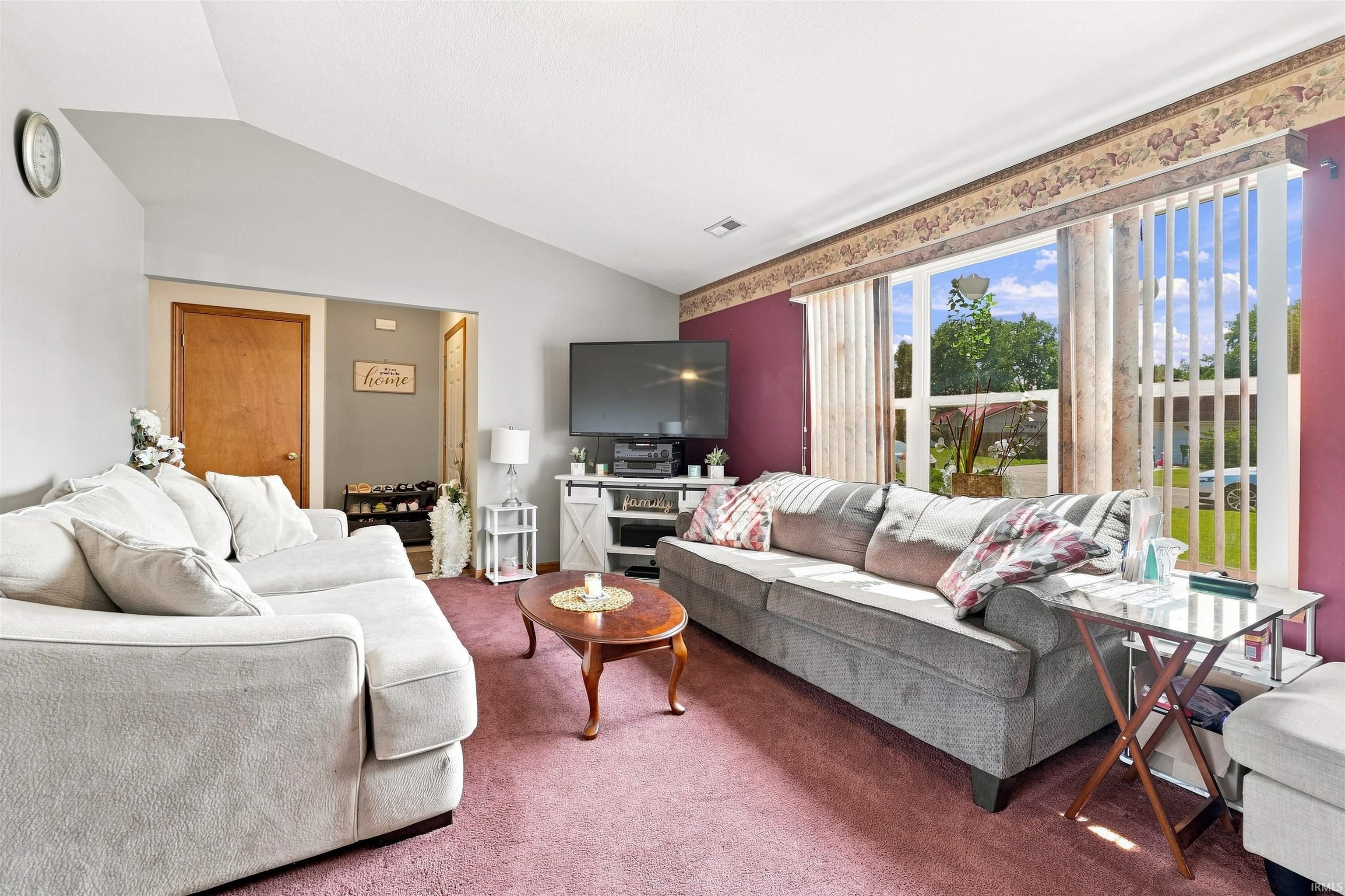 Spacious living area featuring a vaulted ceiling, large picture window with vertical blinds, and plush carpeting