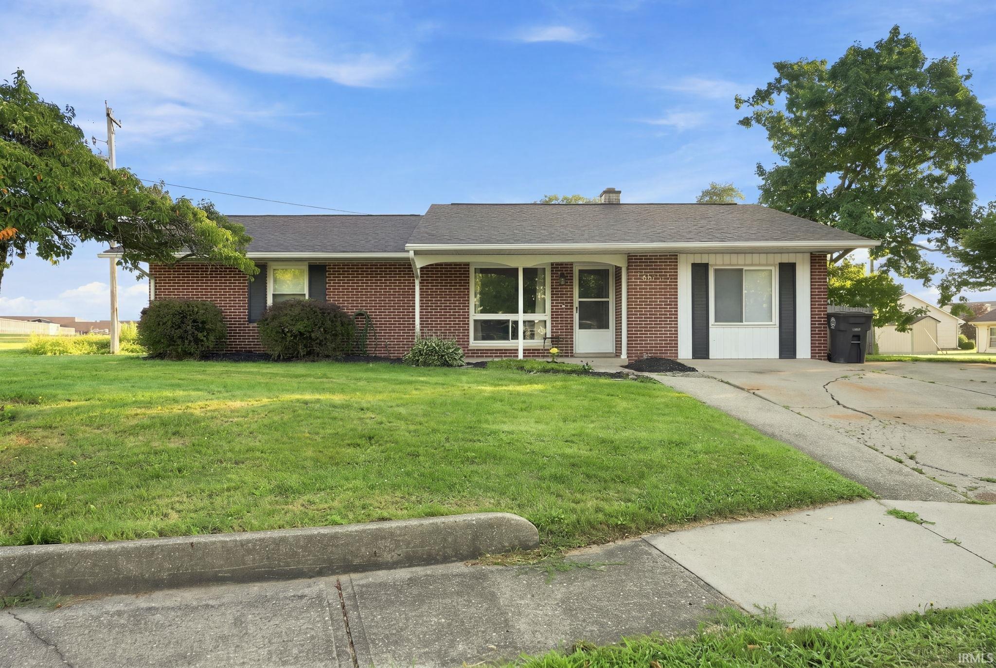 Brick ranch-style home featuring a covered entry, established landscaping, a concrete driveway, and a front lawn with mature trees