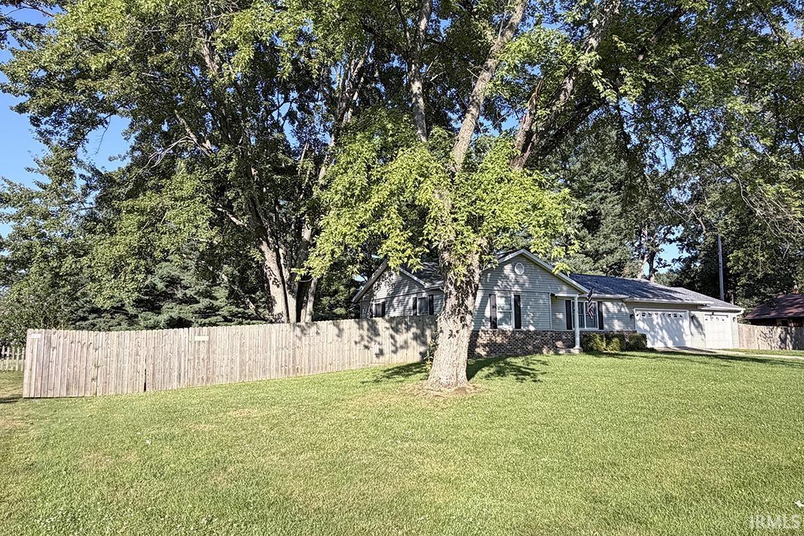 Front exterior featuring a light gray siding, stone accent facade, attached two-car garage, mature trees, and a wooden privacy fence