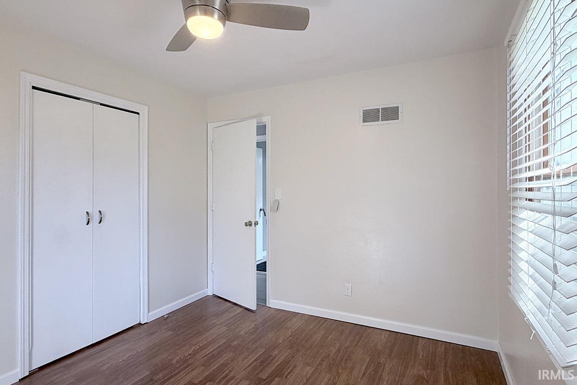 Spacious bedroom featuring wood-finish flooring, a ceiling fan with integrated lighting, dual closets with white frame doors, and large windows with plantation shutters providing natural light and privacy