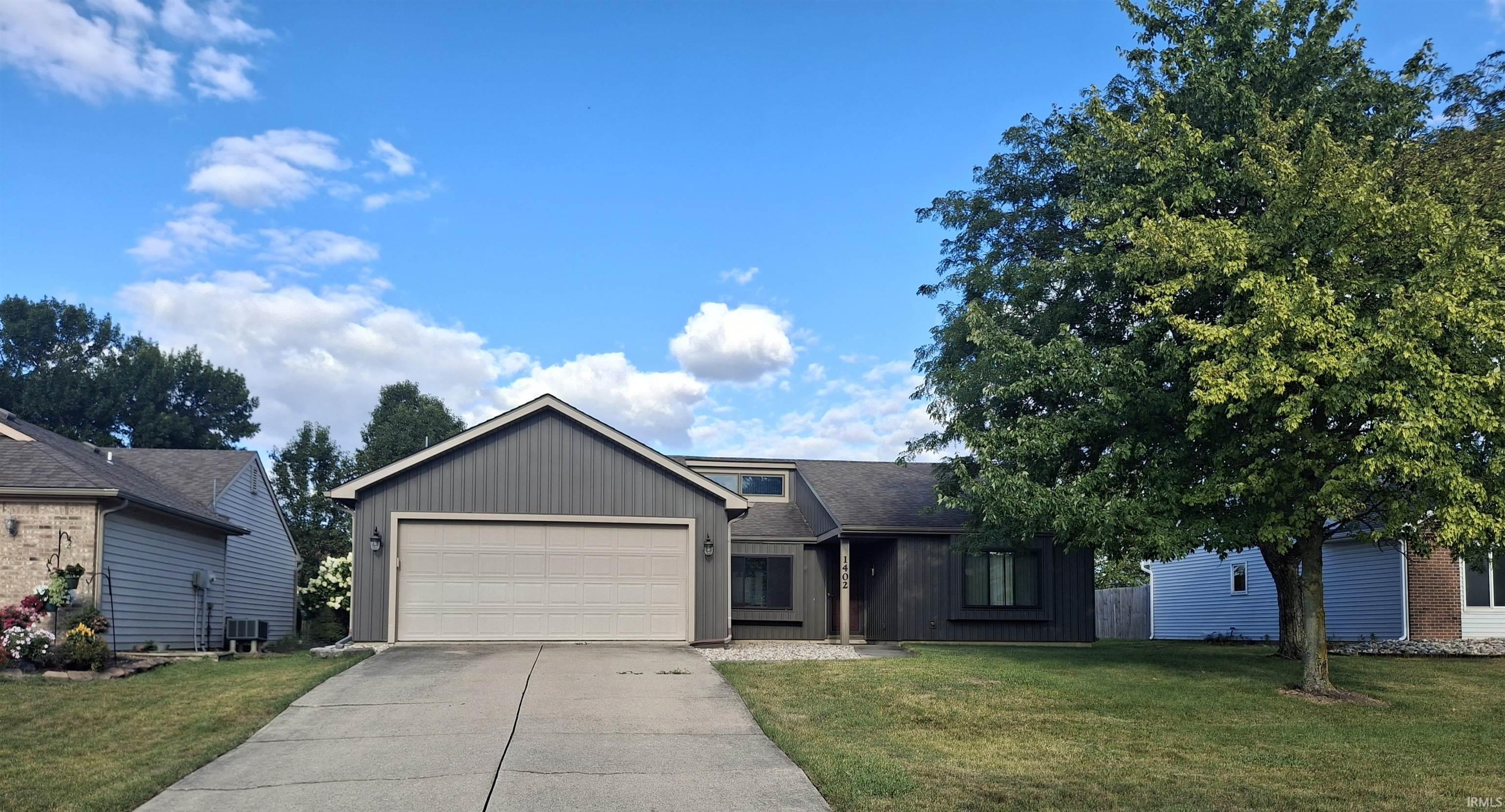Contemporary residence featuring a two-car garage with a light-colored door, dark gray vertical and horizontal siding, a prominent gable roofline, and a mature front yard tree