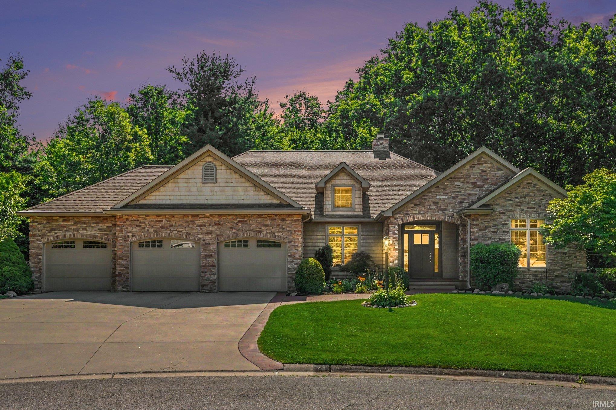 Brick and stone exterior with a three-car garage, gabled roofline, and professionally landscaped front grounds featuring mature trees and a manicured lawn