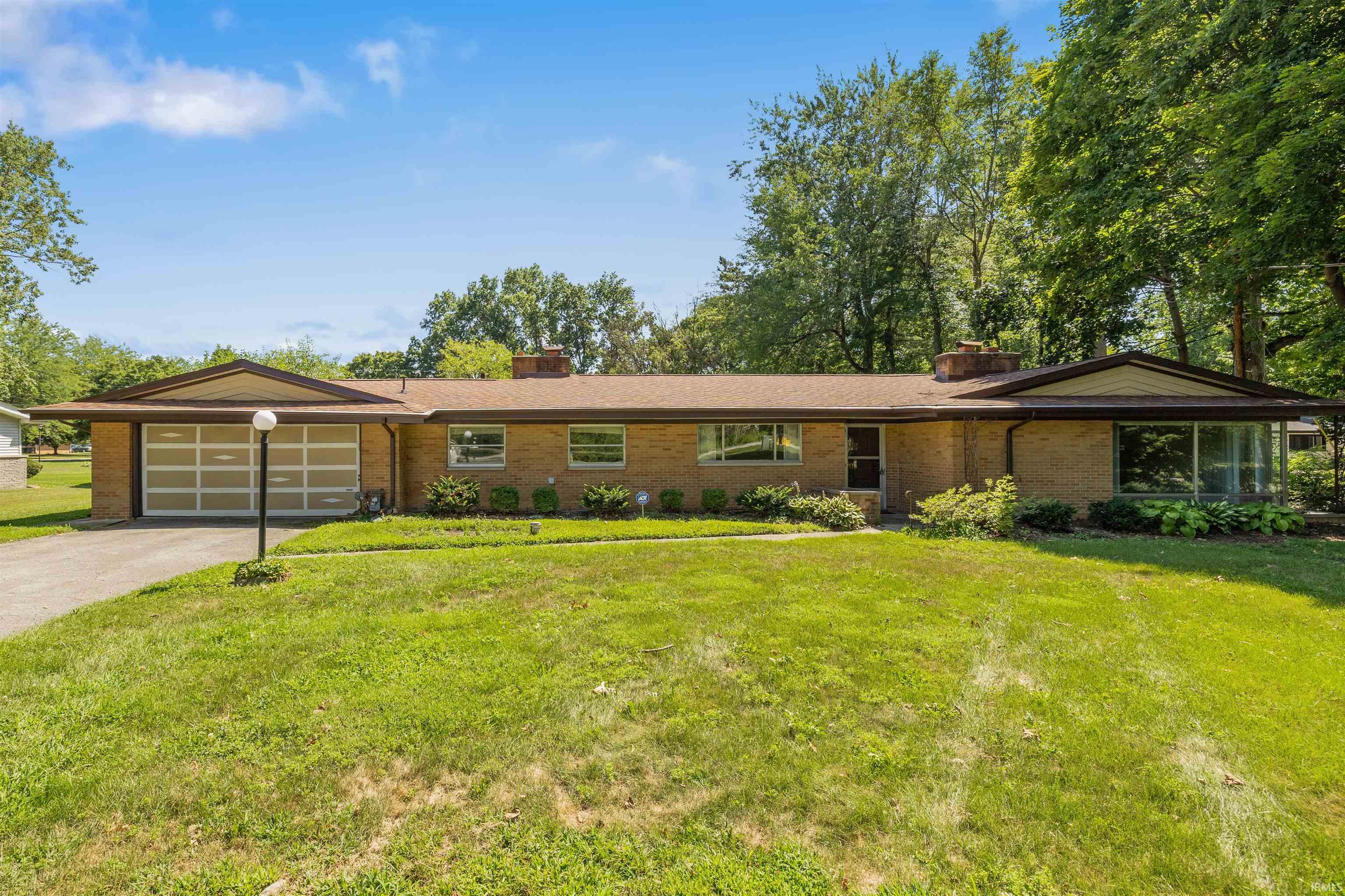 Mid-century brick ranch featuring a two-car attached garage with modern paneled doors, expansive front lawn with mature landscaping, and a low-pitched roofline characteristic of classic residential architecture
