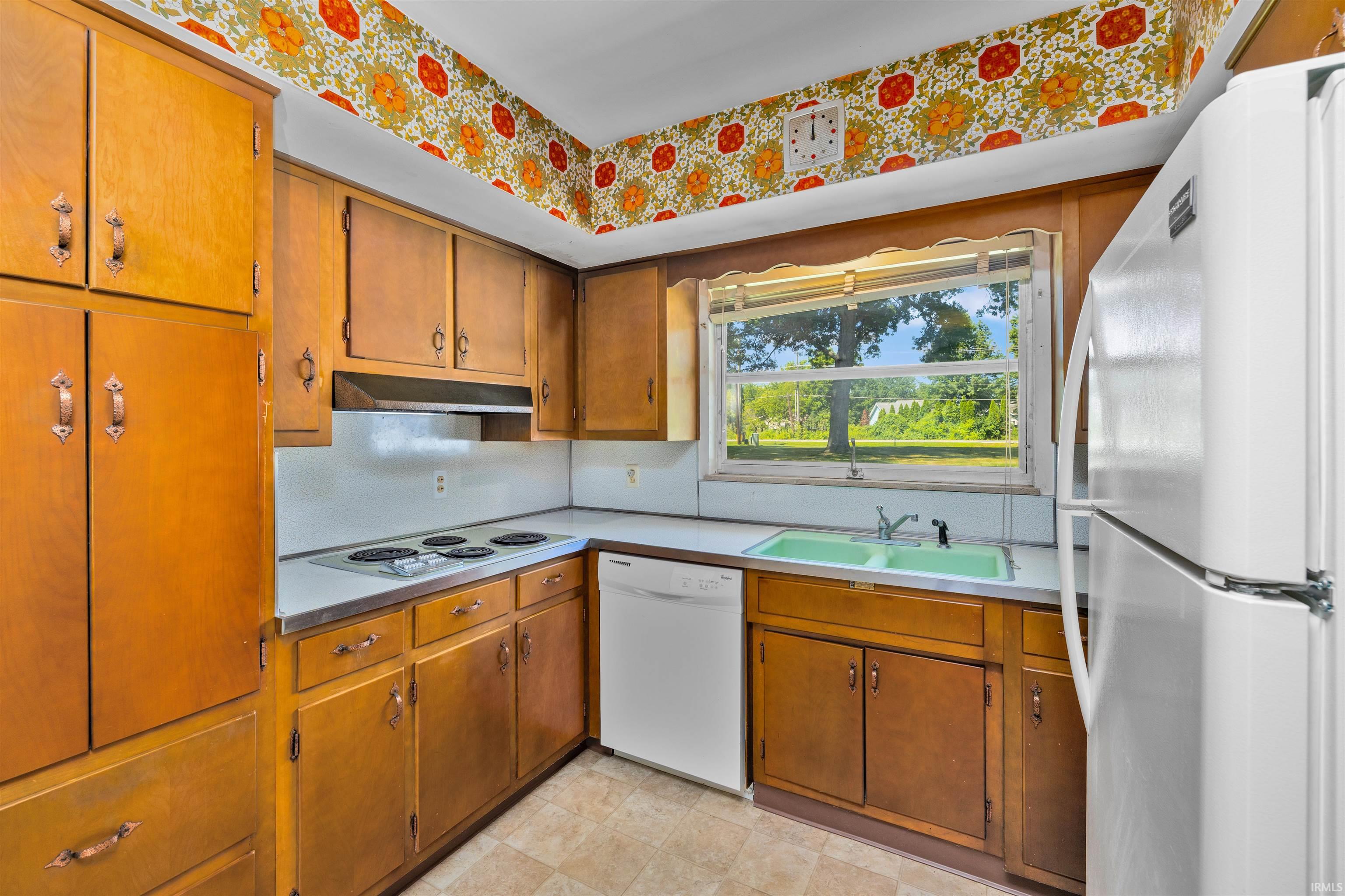 Charming kitchen featuring warm wood cabinetry, white appliances including a refrigerator and electric range, stainless steel sink with window overlooking outdoor scenery, and retro floral wallpaper detailing the ceiling