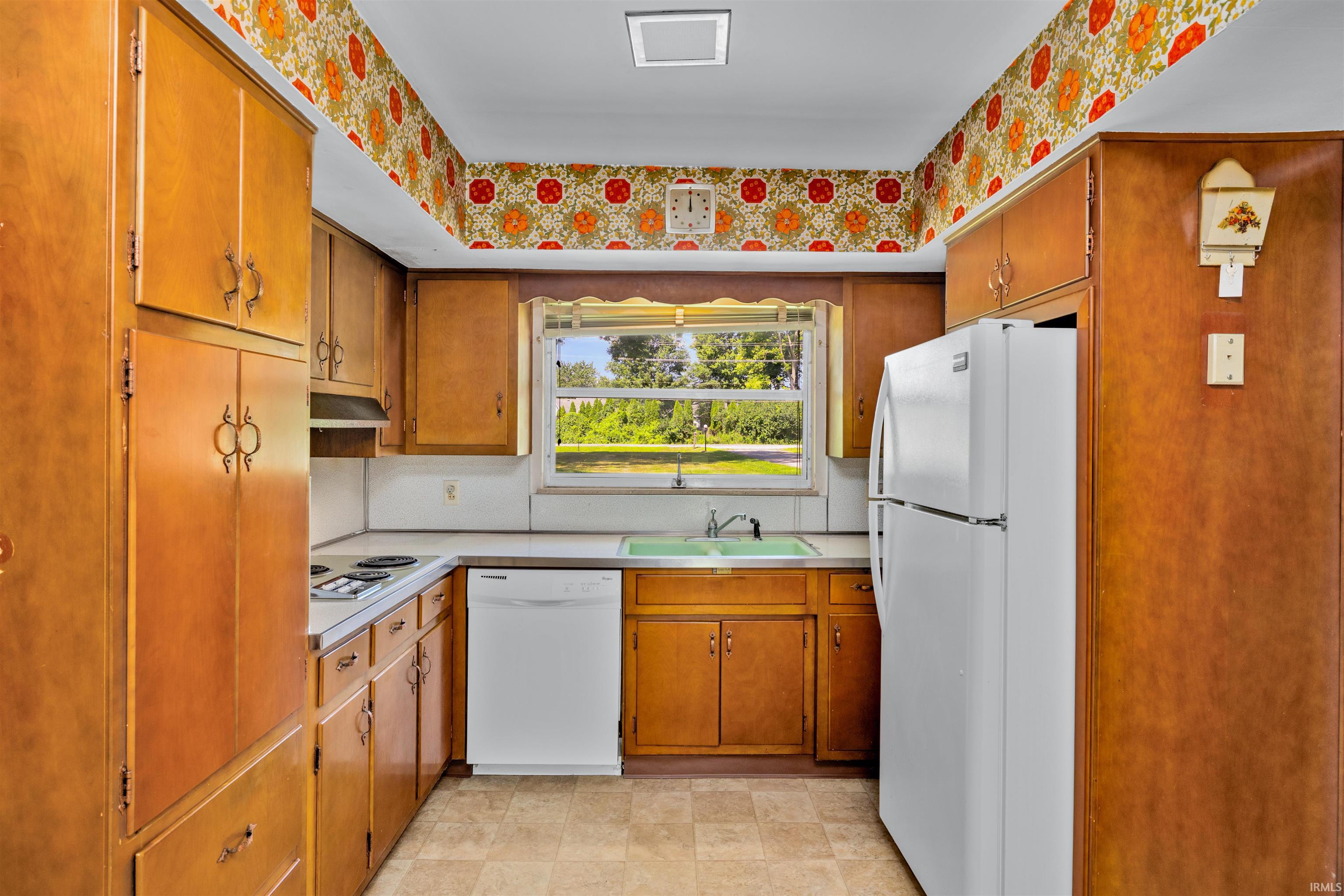 Galley kitchen featuring warm wood cabinetry, white appliances including a refrigerator and dishwasher, laminate countertops, and a window overlooking outdoor scenery