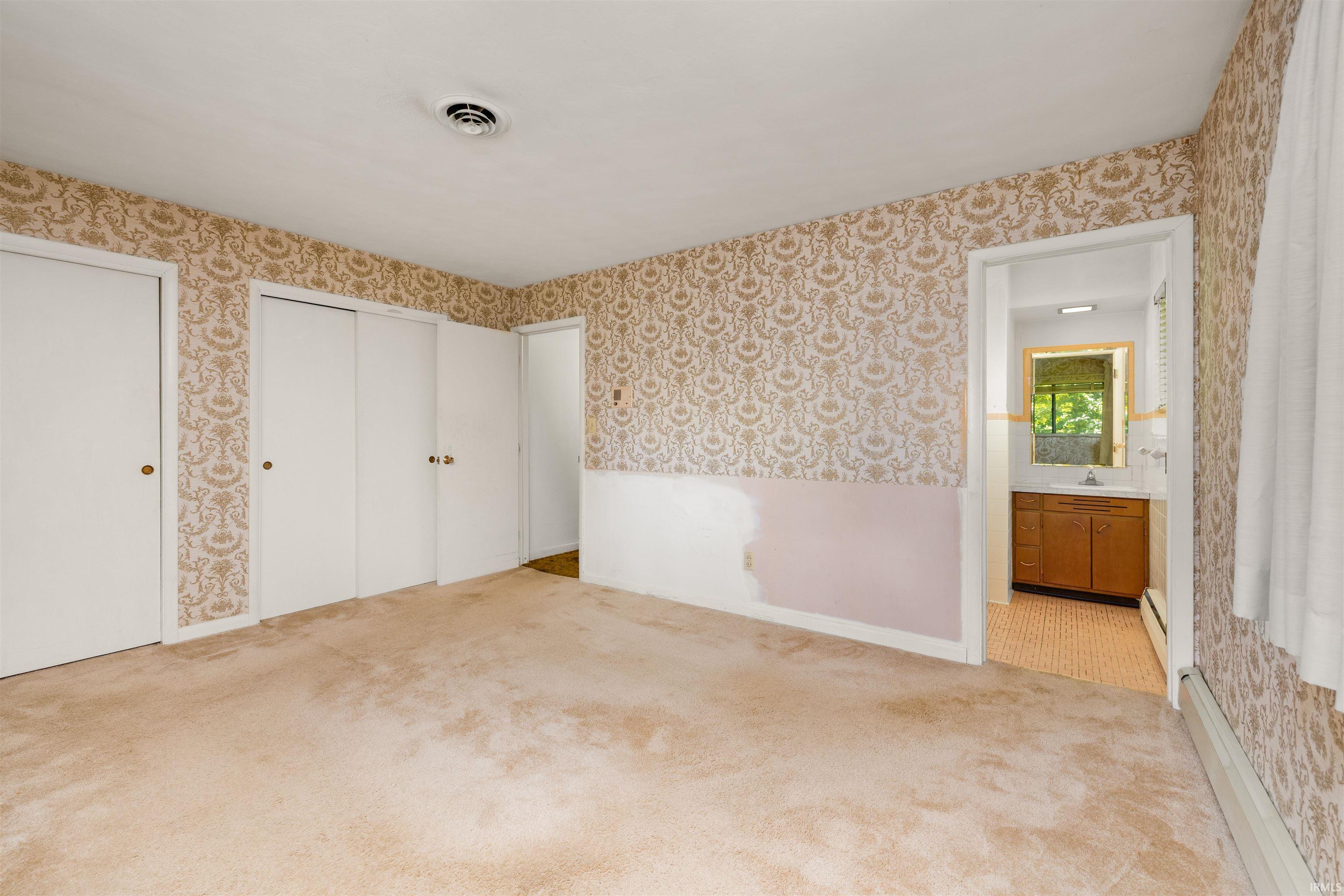 Spacious bedroom featuring ornamental patterned wallcovering, white panel closet doors with brass hardware, neutral wood-finish flooring, and recessed ceiling lighting