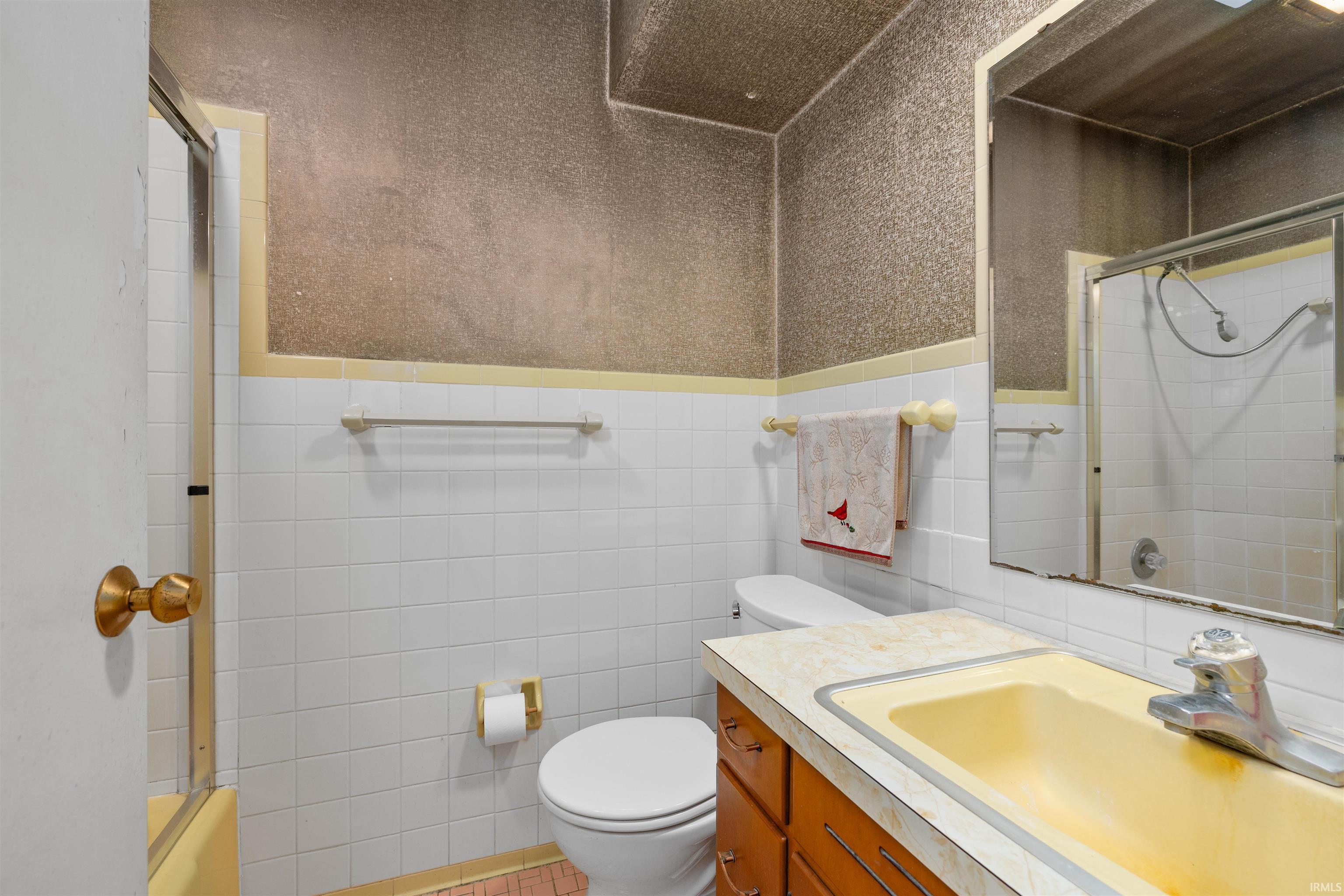 Bathroom featuring white subway tile wainscoting with textured upper walls, wood-finish vanity cabinetry, dual sink countertop with yellow laminate surface, framed mirror with brass-tone hardware, and coordinating brass towel bars