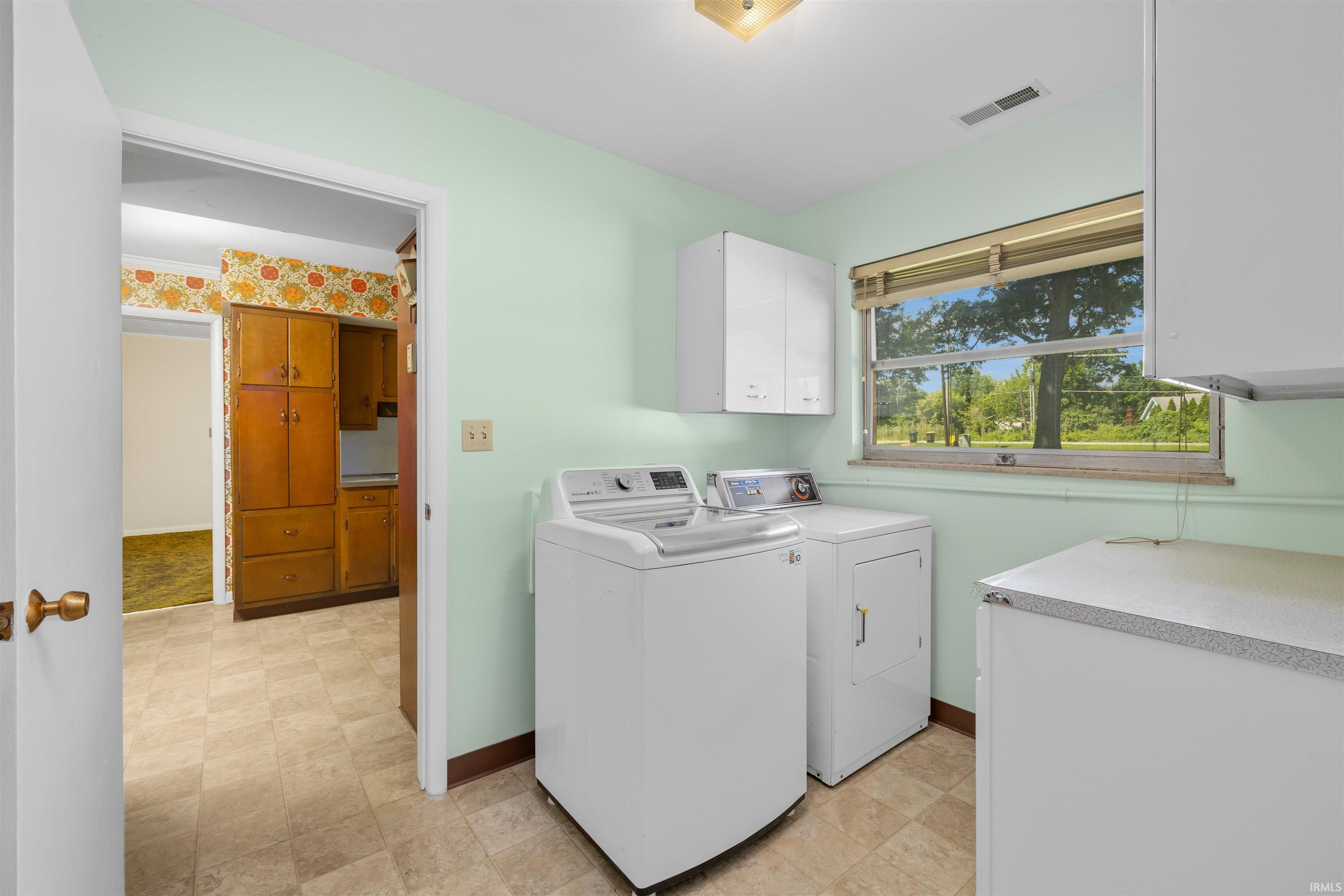 Laundry area featuring light green walls, a window overlooking exterior greenery, two upper white cabinets, and durable tile flooring