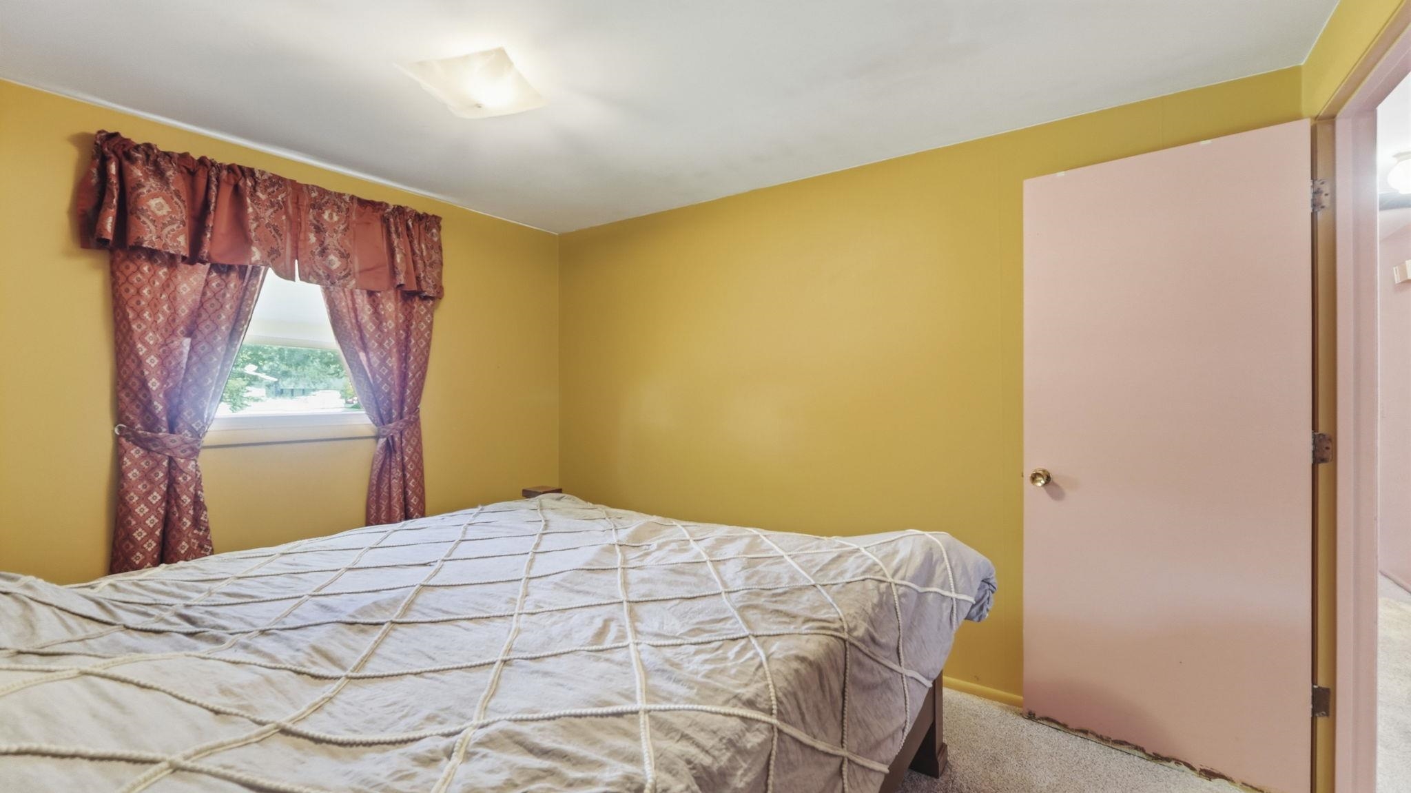 Bright and cheerful bedroom featuring soft yellow walls, carpeted flooring, and a large window with burgundy valance and curtains