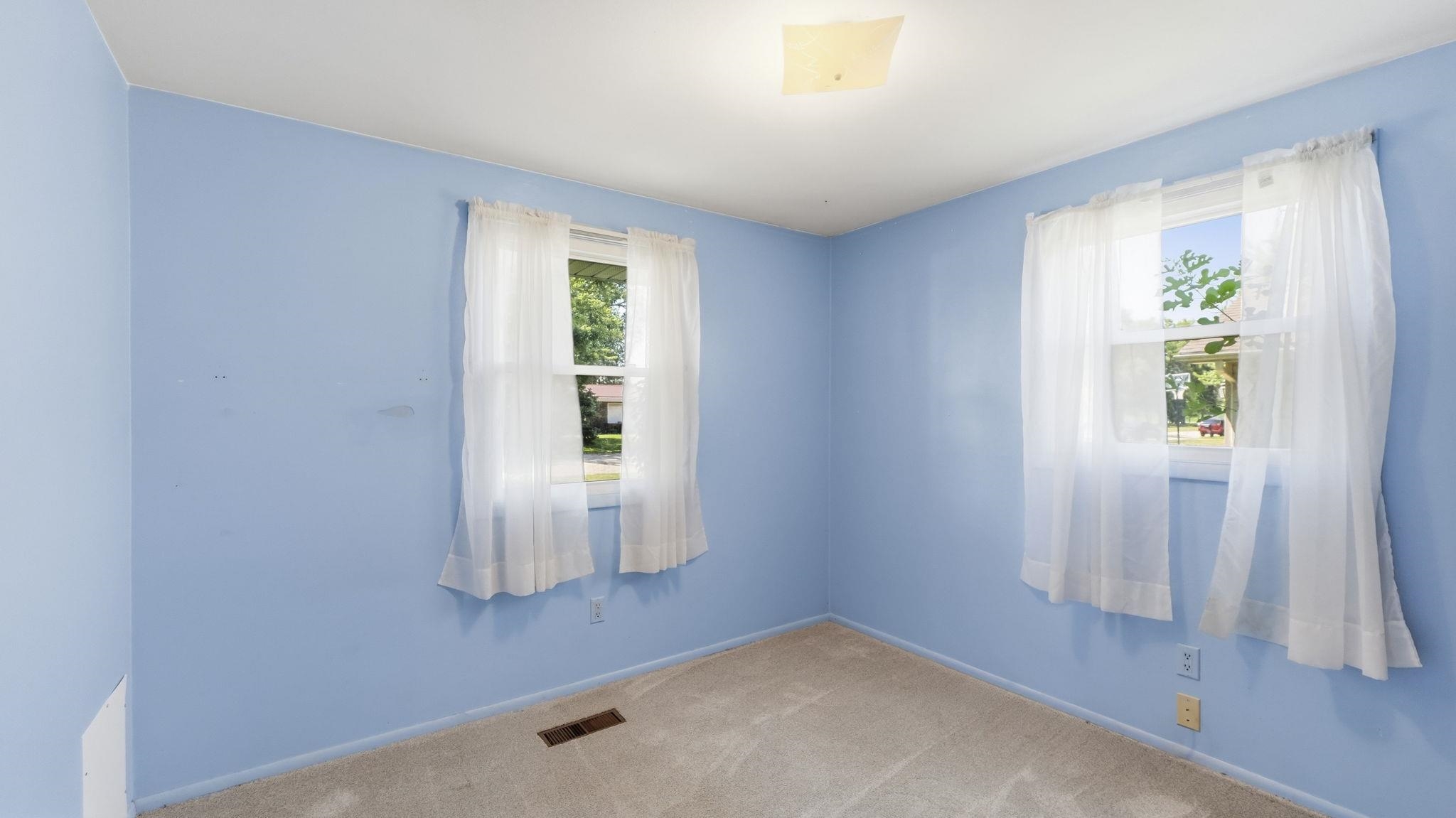 Bright secondary bedroom featuring periwinkle walls, dual windows with sheer curtains allowing natural light, and neutral tile flooring