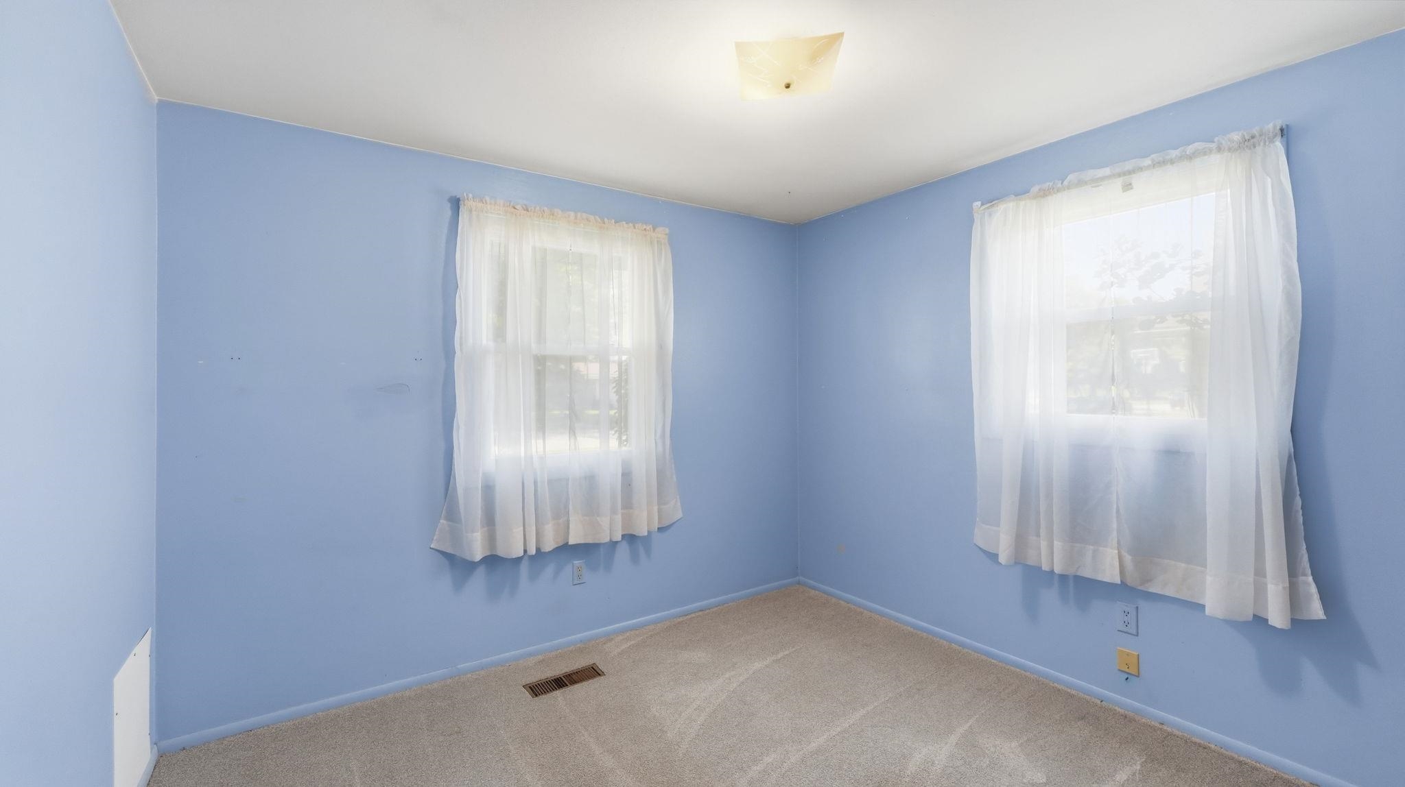 Bright secondary bedroom featuring periwinkle walls, wood-finish flooring, and dual windows with sheer curtains providing natural light throughout the space