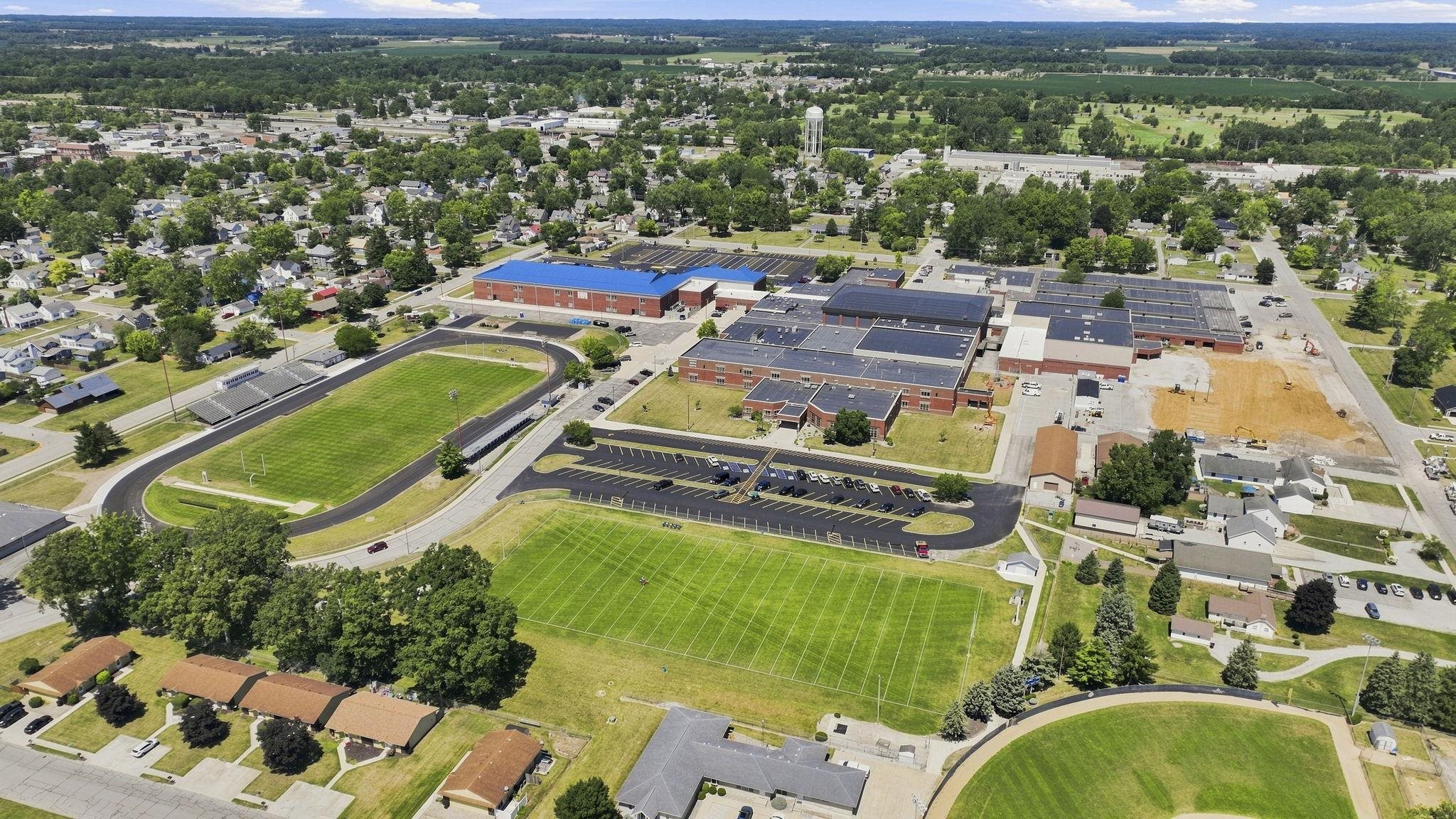 Aerial view showcasing this property's expansive grounds featuring a regulation-size athletic track, multiple sports fields with manicured turf, modern recreational facilities with blue roof paneling, and substantial paved areas
