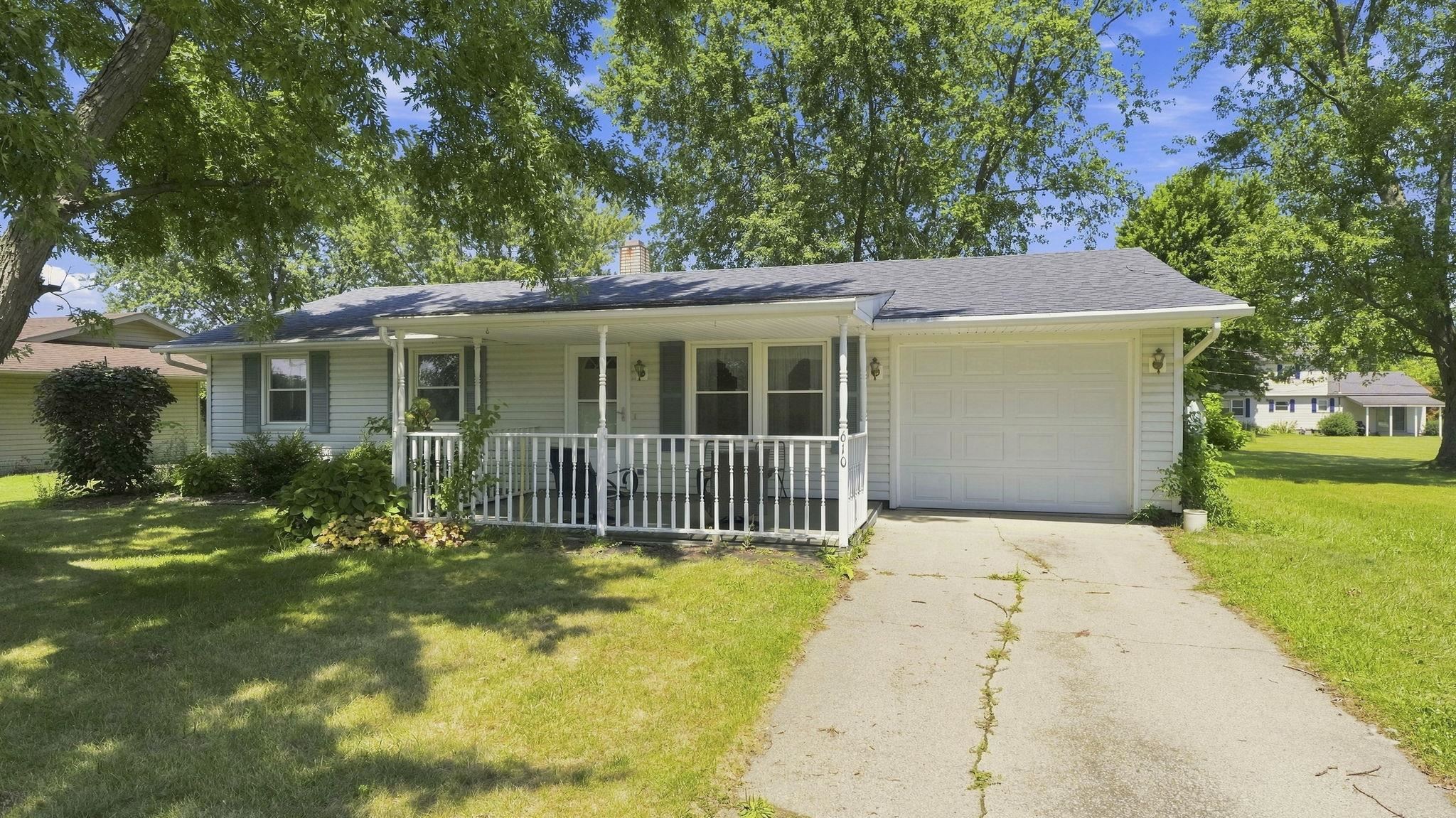 Charming ranch home featuring a welcoming front porch with white railings, attached two-car garage, and mature landscaping with established trees providing natural shade