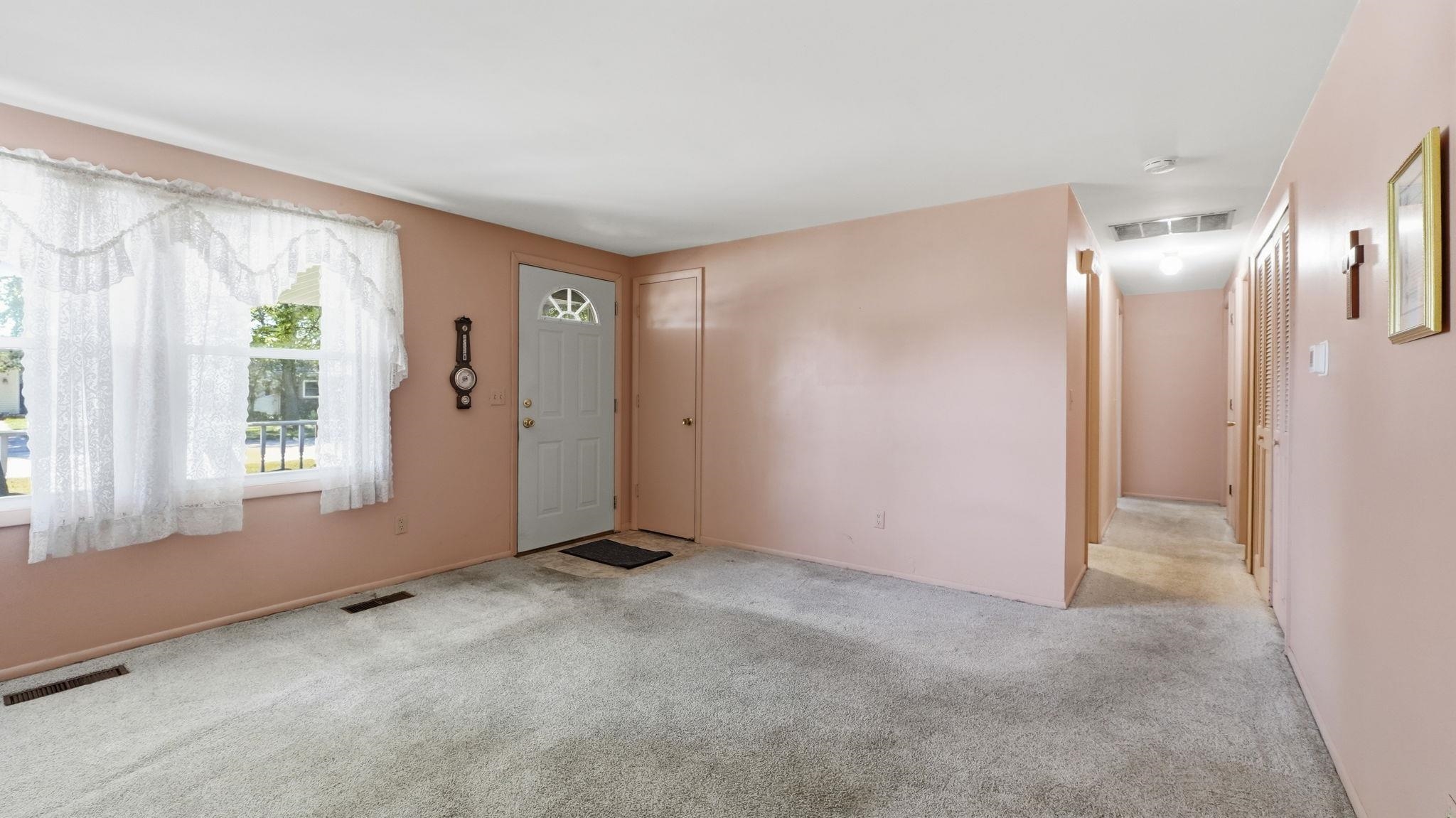 Spacious living area showcasing neutral pink walls, plush carpet flooring, and abundant natural light from a front-facing window