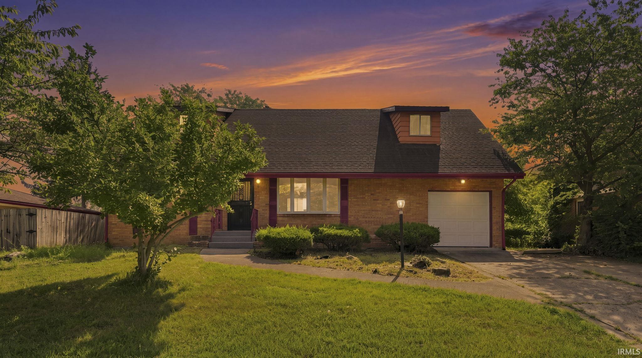 Brick exterior featuring a dormer window, an attached garage with a white door, a paved driveway, and mature landscaping