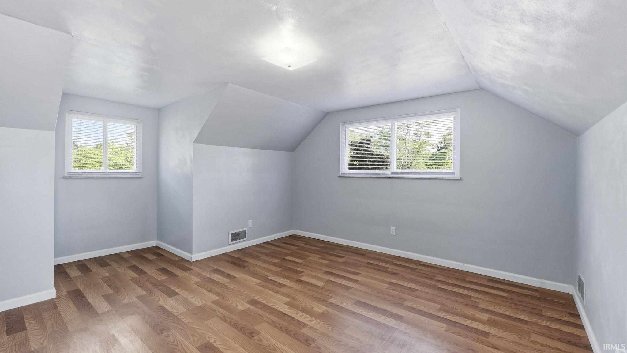 Room featuring wood-finish flooring, two windows, vaulted ceiling sections, light blue wall paint, and a ceiling-mounted light fixture