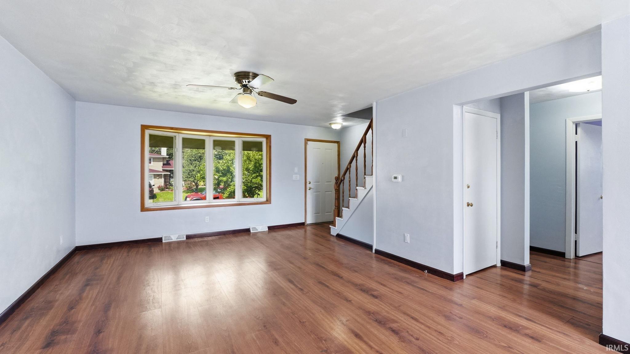 Spacious open-concept room featuring wood-finish flooring, a large bay window with wood trim, a ceiling fan with integrated lighting, and a staircase with wood railings