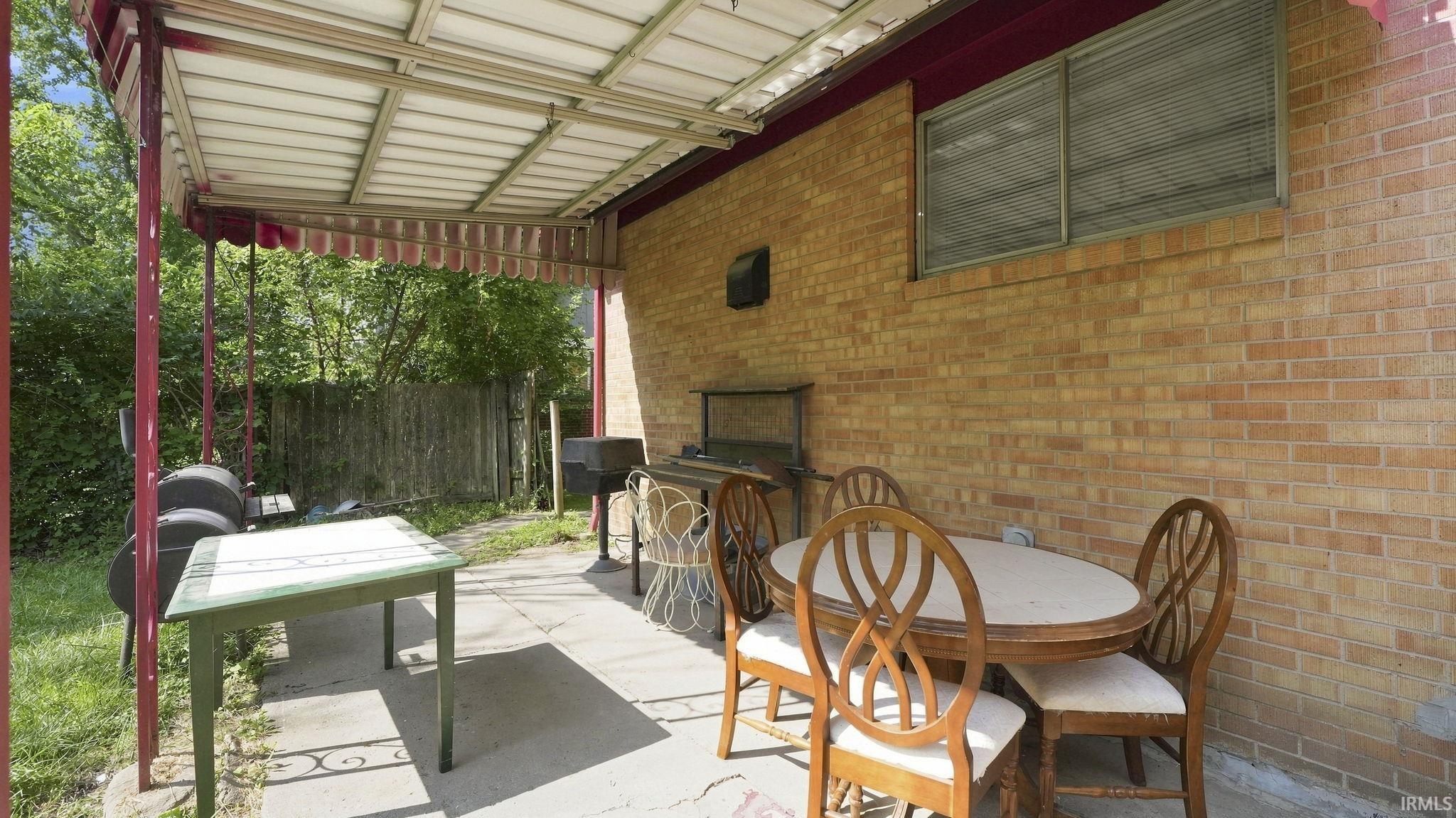Covered concrete patio featuring a corrugated metal awning with red trim, brick exterior wall, and a horizontal sliding window