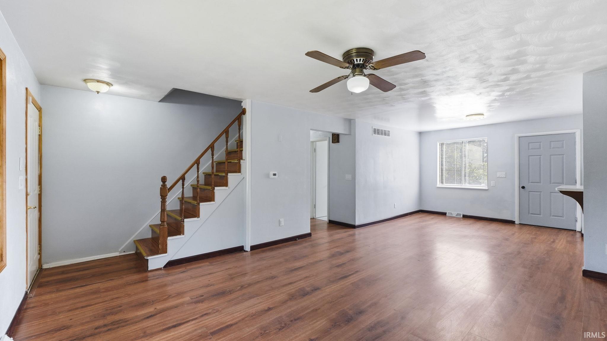 Spacious room featuring wood-finish flooring, a ceiling fan with integrated lighting, and a prominent staircase with wood handrails