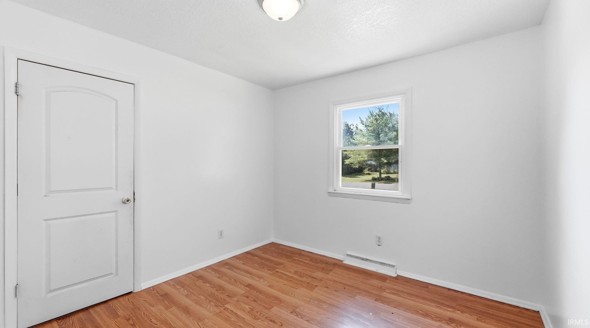 Room featuring wood-finish flooring, a white interior door with an arched panel, a window with white trim, and a ceiling-mounted light fixture