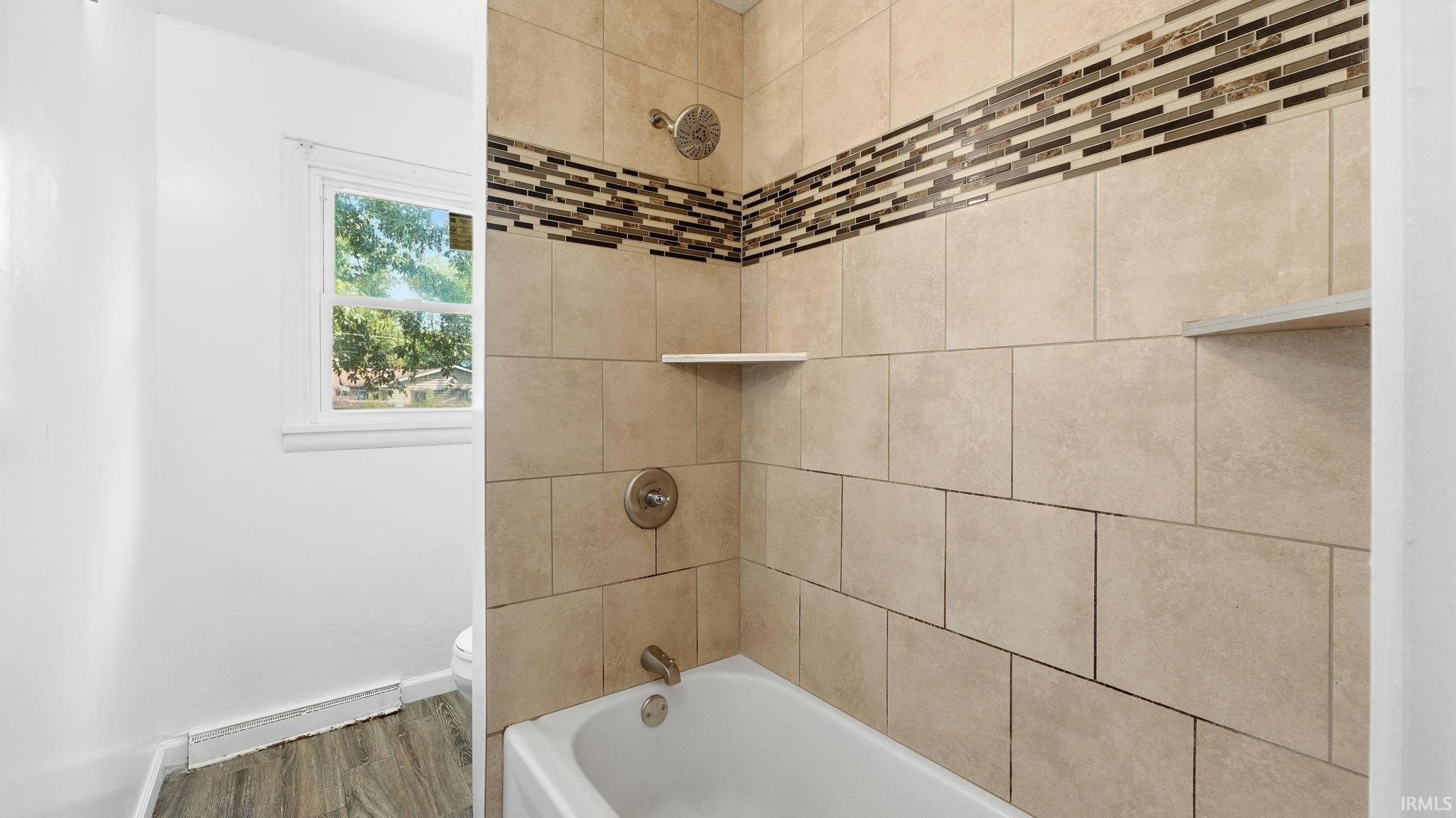 Bathtub and shower combination featuring neutral-toned tile surrounds, a decorative mosaic tile accent band, and integrated shelving