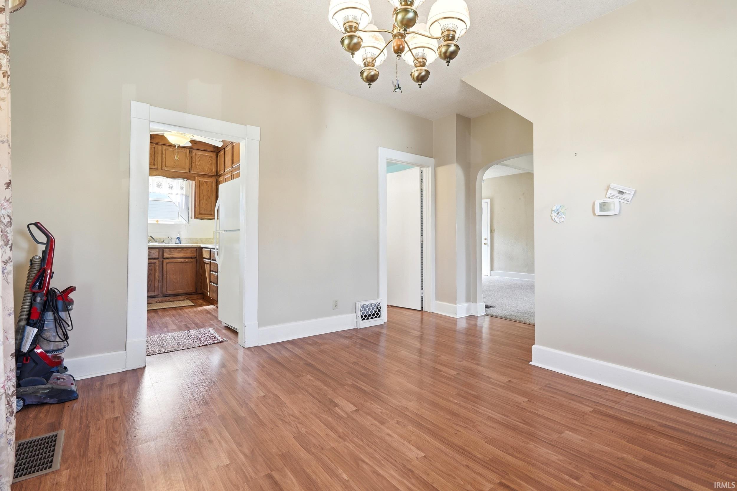 Wood-finish flooring throughout the main area with white baseboards, a brass chandelier, and an arched entryway to an additional room