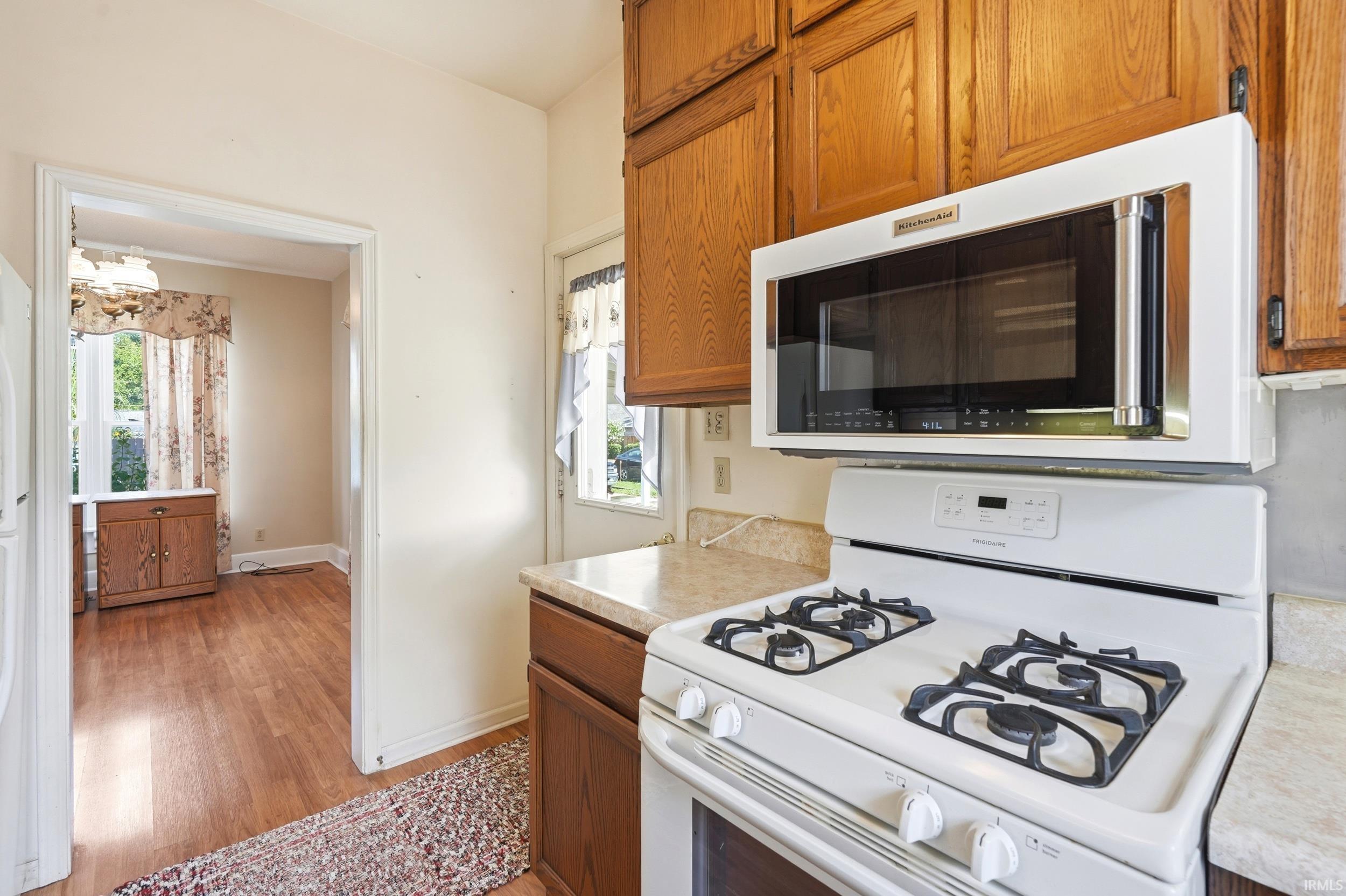 Kitchen featuring wood cabinetry, white appliances, laminate countertops, and wood-finish flooring extending into an adjacent room