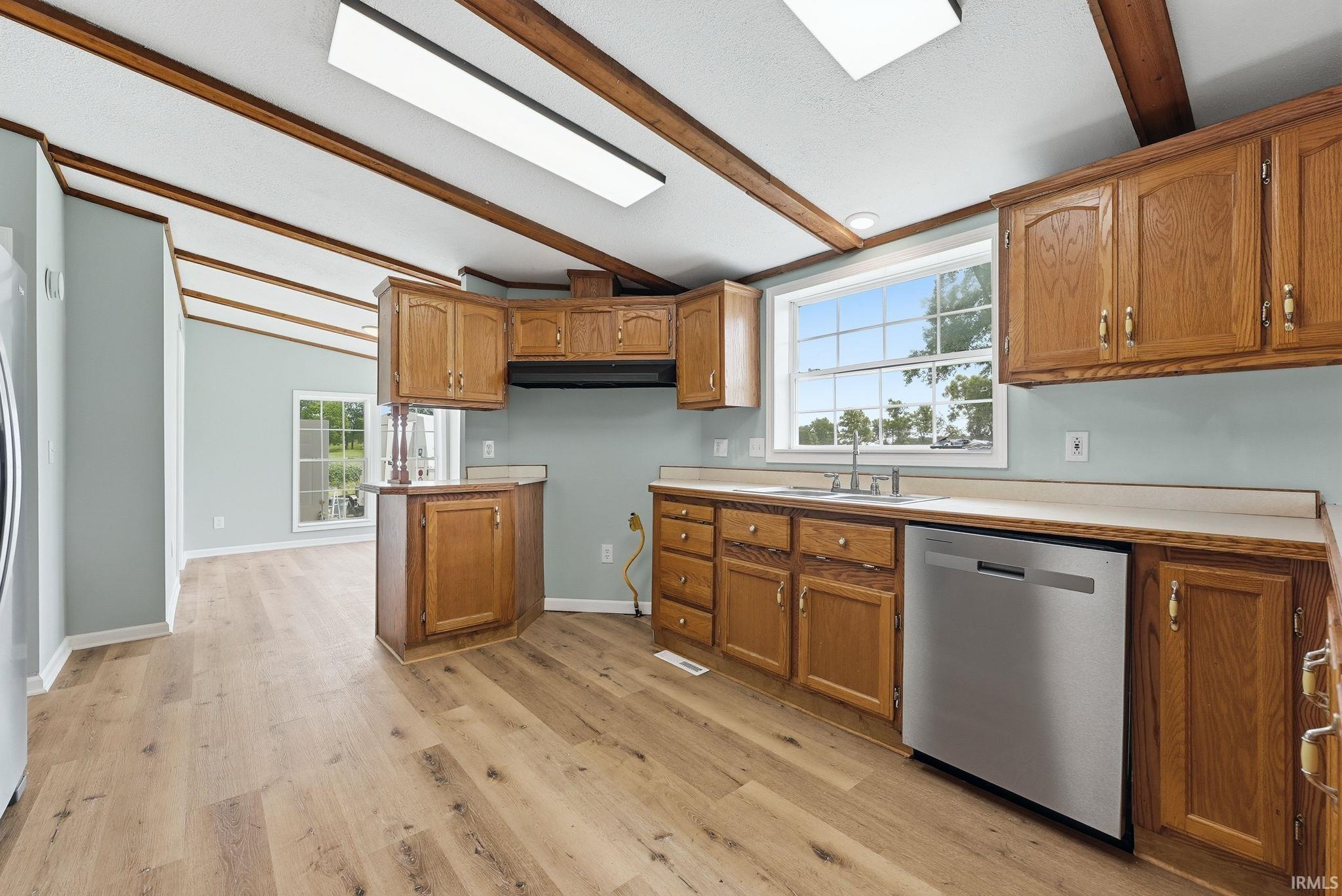 Kitchen featuring wood-finish flooring, wood cabinetry with gold-tone hardware, a stainless steel dishwasher, and a double basin sink beneath a window