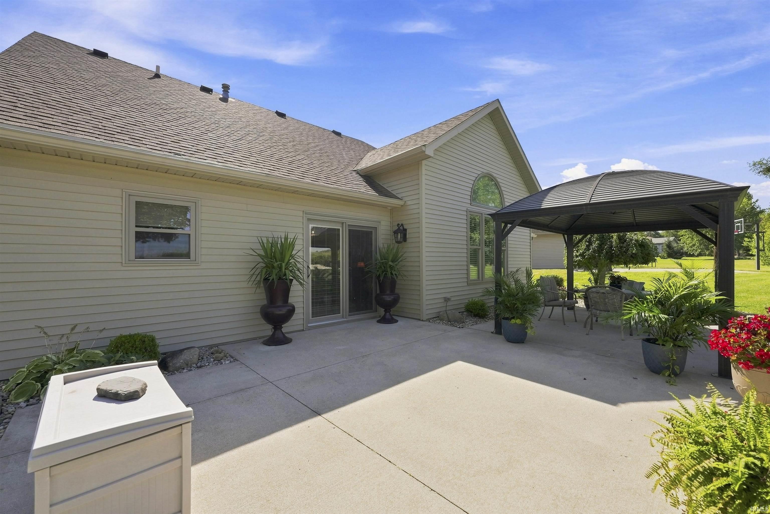 Inviting rear patio featuring a spacious concrete entertaining area, attached pergola structure with integrated shade, manicured landscaping with seasonal plantings, and expansive lawn backed by mature trees