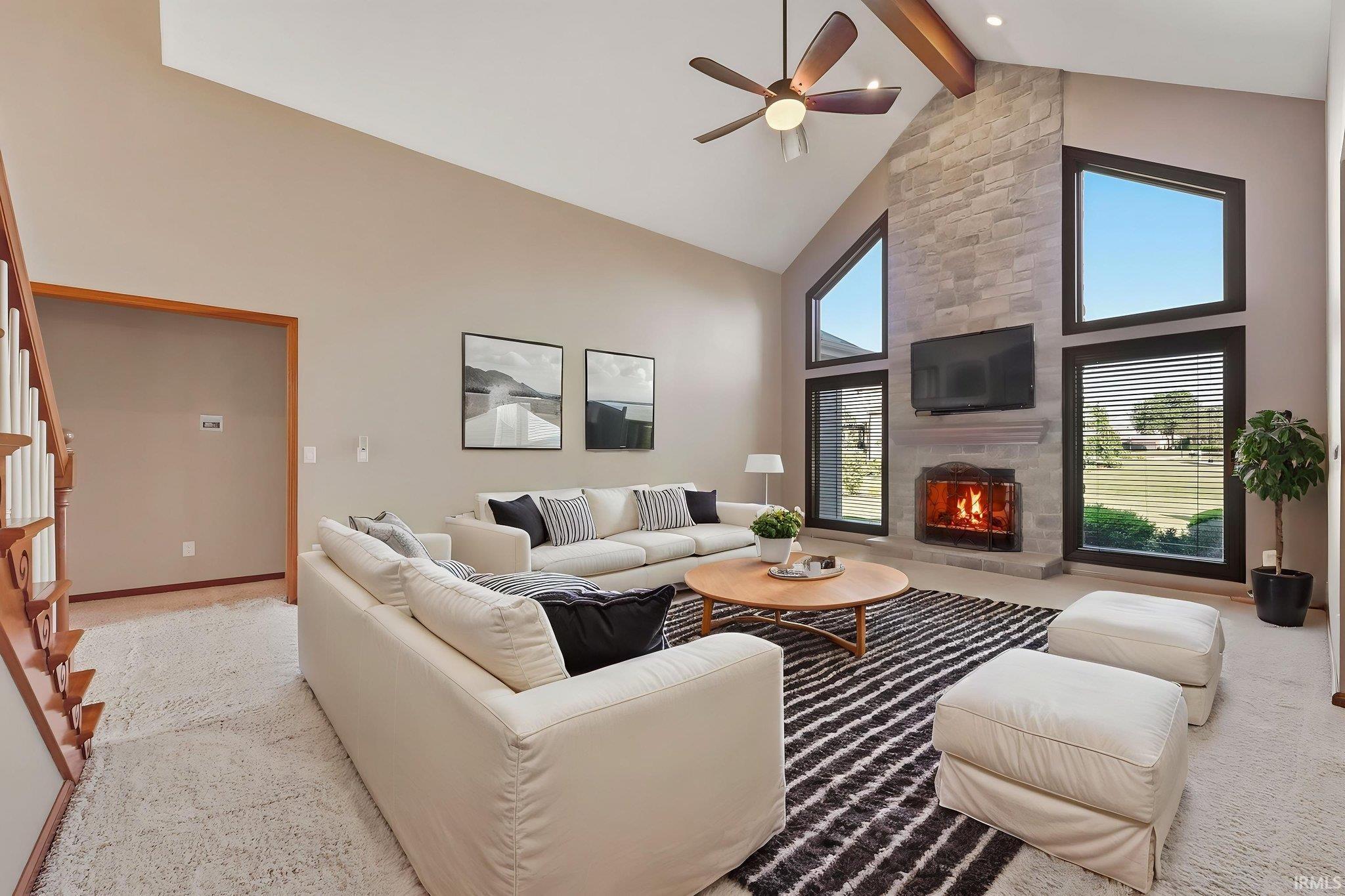 Vaulted ceiling living room featuring a floor-to-ceiling stone-clad fireplace, large picture windows with dark trim, a wood-finish ceiling fan, and carpet flooring