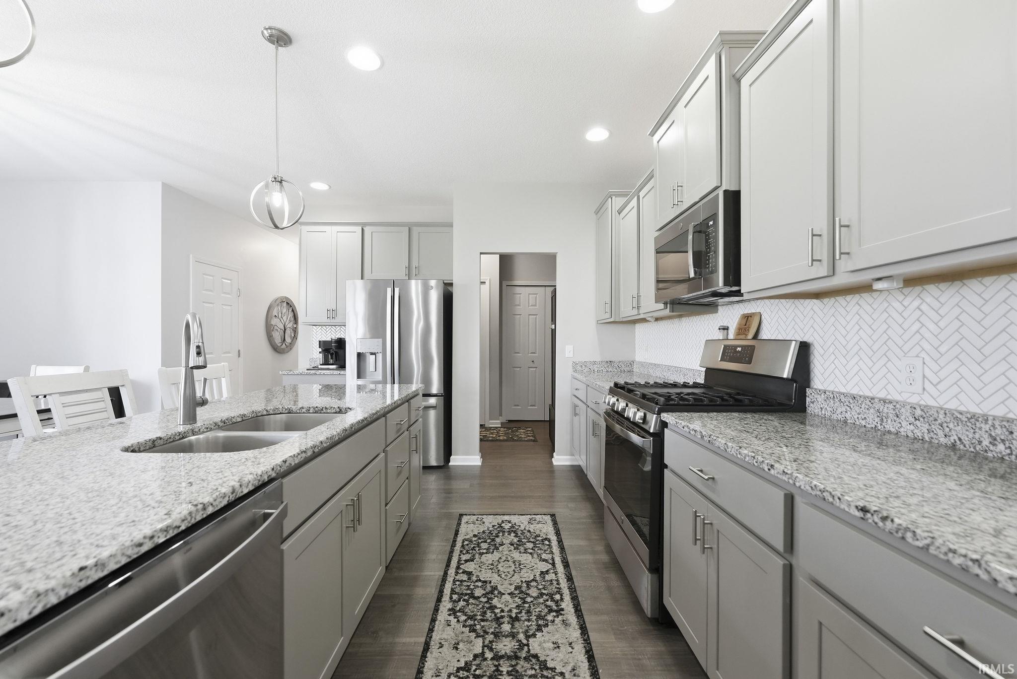 Modern kitchen featuring stainless steel appliances, granite countertops with waterfall edge detail, white cabinetry with brushed nickel hardware, herringbone tile backsplash, pendant lighting, and dark wood-finish flooring