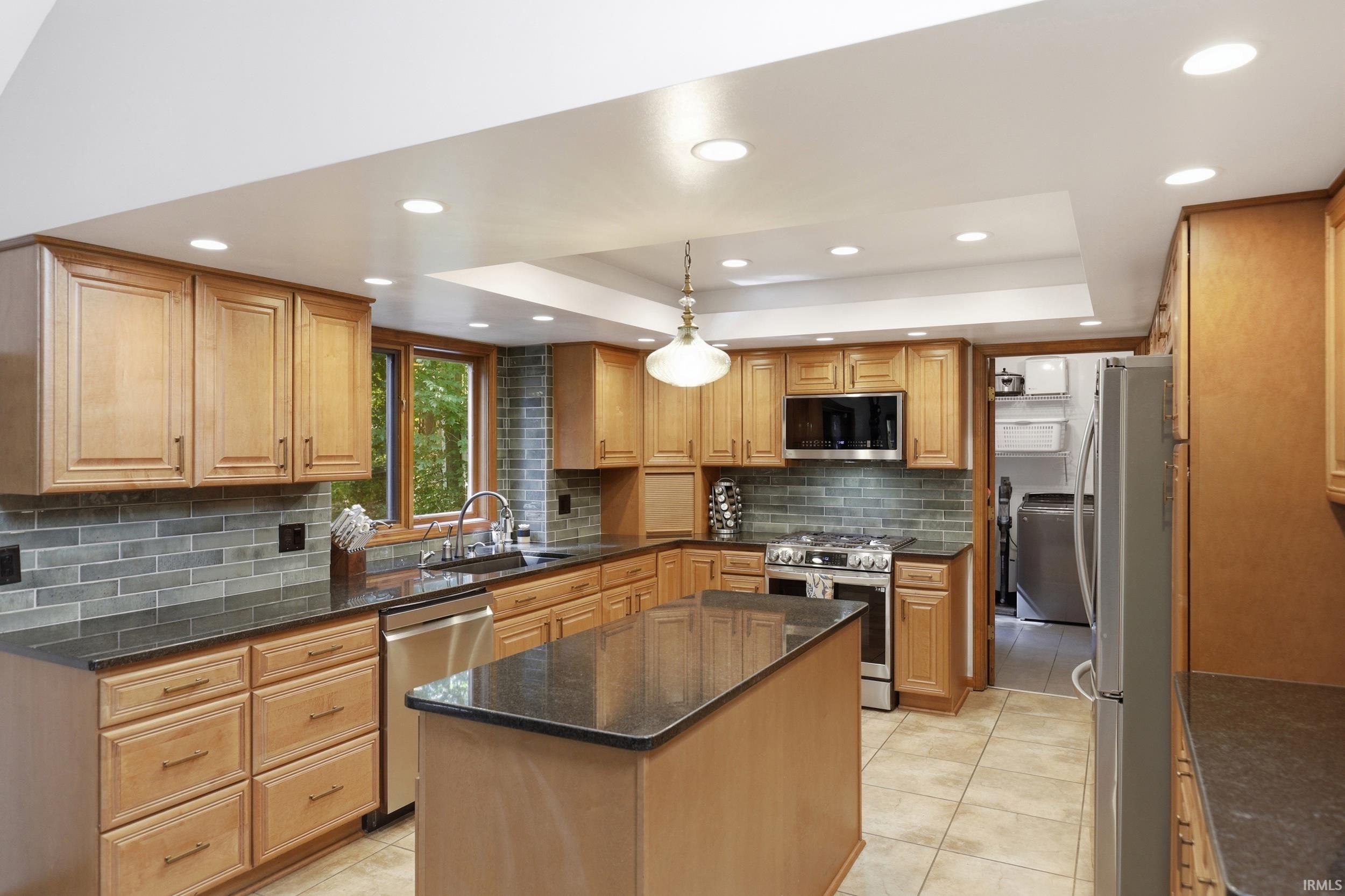 Kitchen featuring light wood cabinetry, dark countertops, a tiled backsplash, stainless steel appliances, and an island with additional counter space