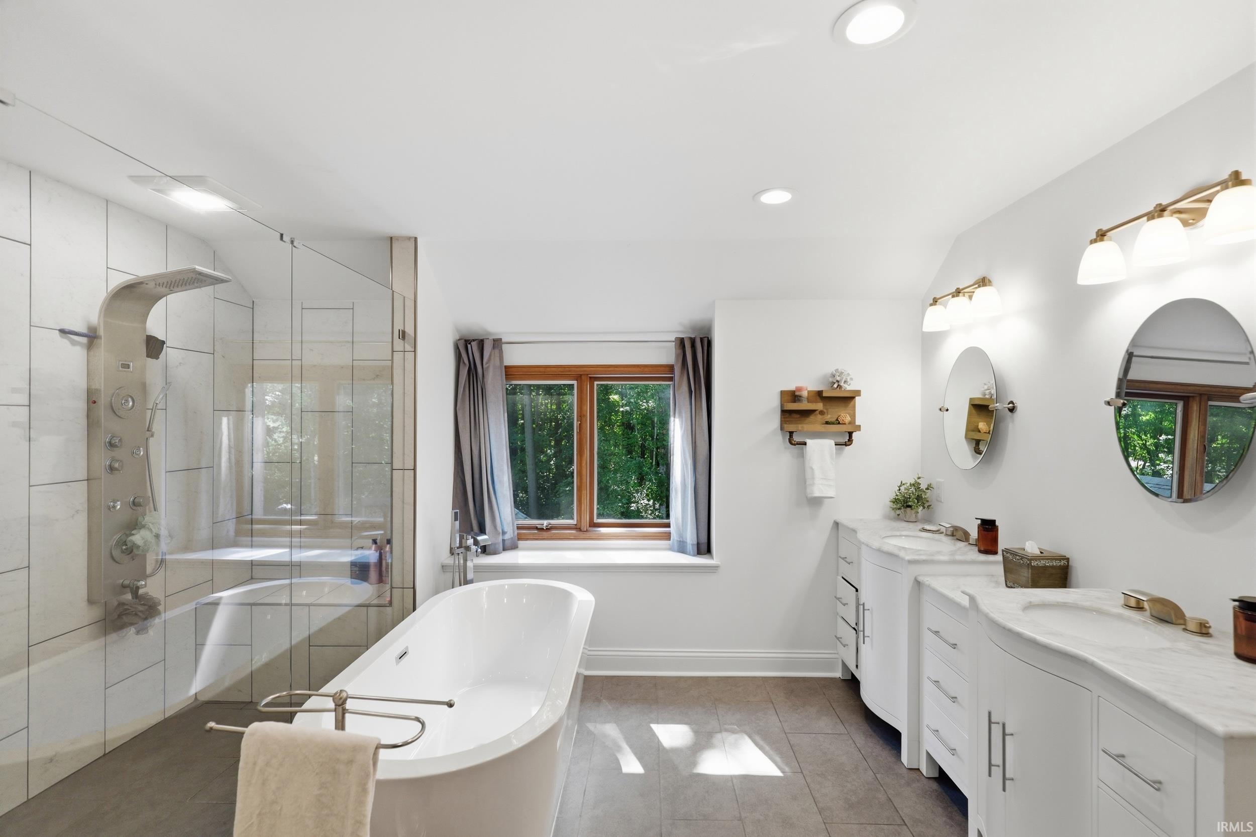 White bathroom featuring a freestanding soaking tub, clear glass shower enclosure with a multi-function shower panel, and a dual vanity with undermount sinks and gold-finish fixtures