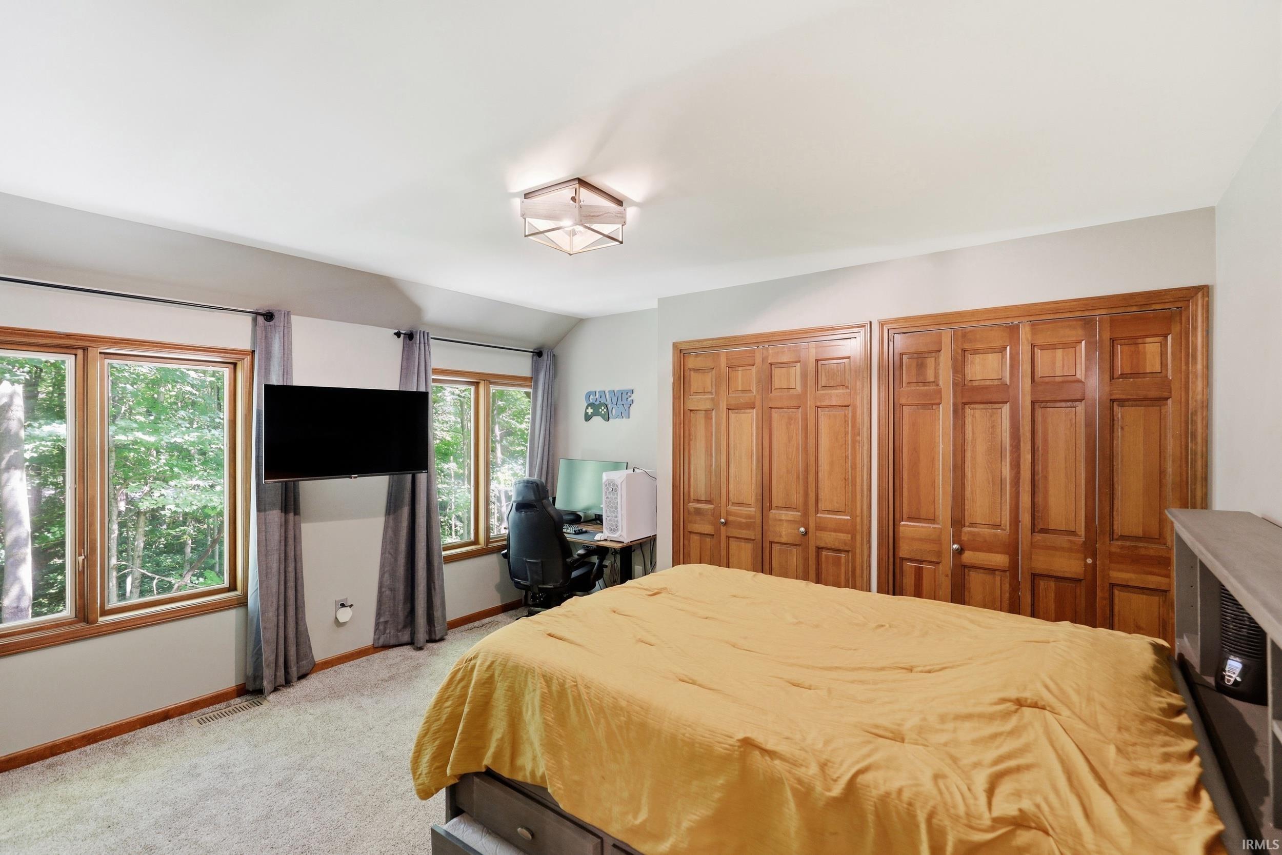 Spacious bedroom featuring multiple windows with wood trim, wall-to-wall carpeting, two sets of bi-fold wood closet doors, and an overhead flush-mount light fixture