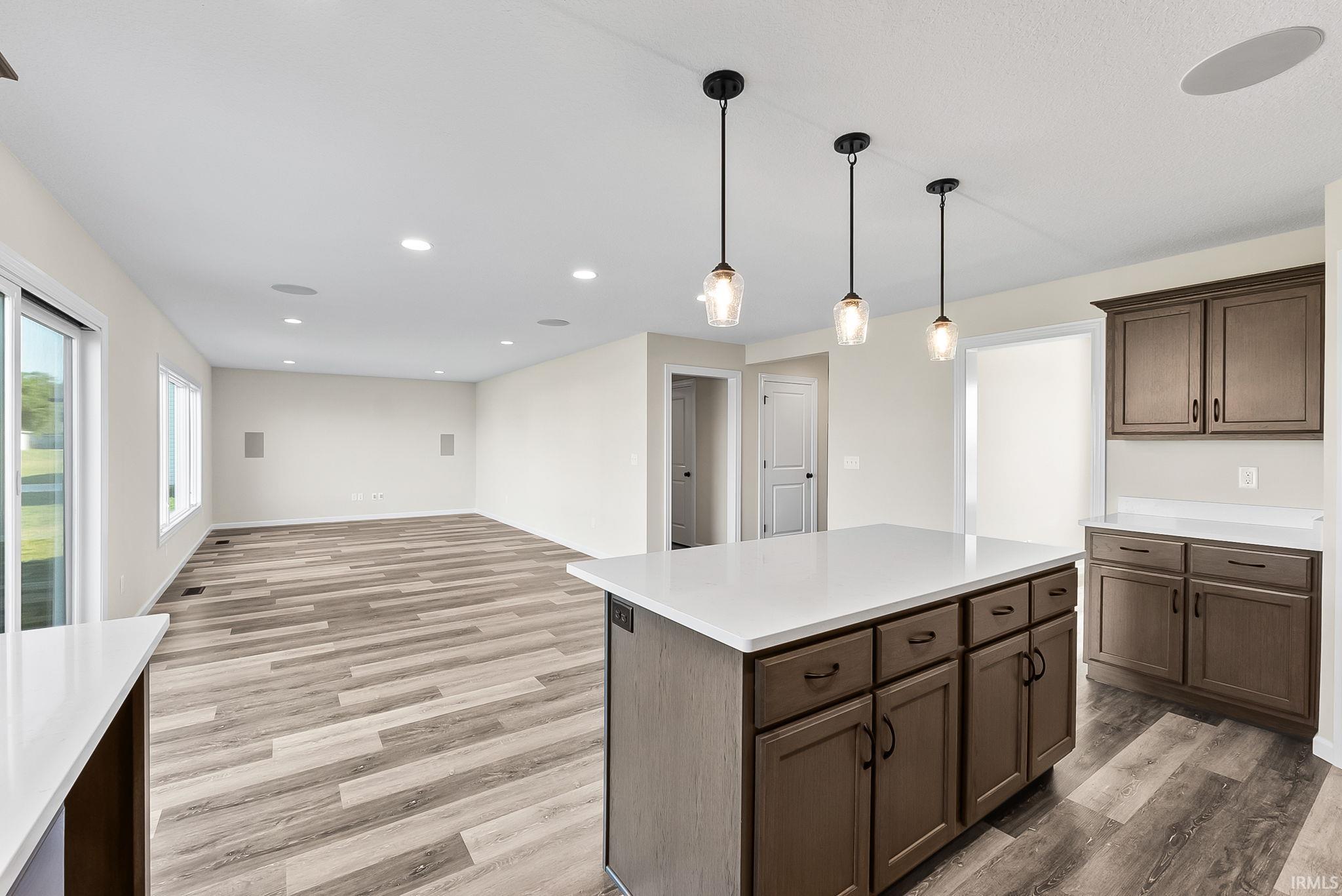 Spacious kitchen featuring a large island with wood cabinetry and drawer storage, white quartz countertop, three pendant lights with glass shades, wood-finish plank flooring, recessed ceiling lighting, and ample natural light from multiple windows