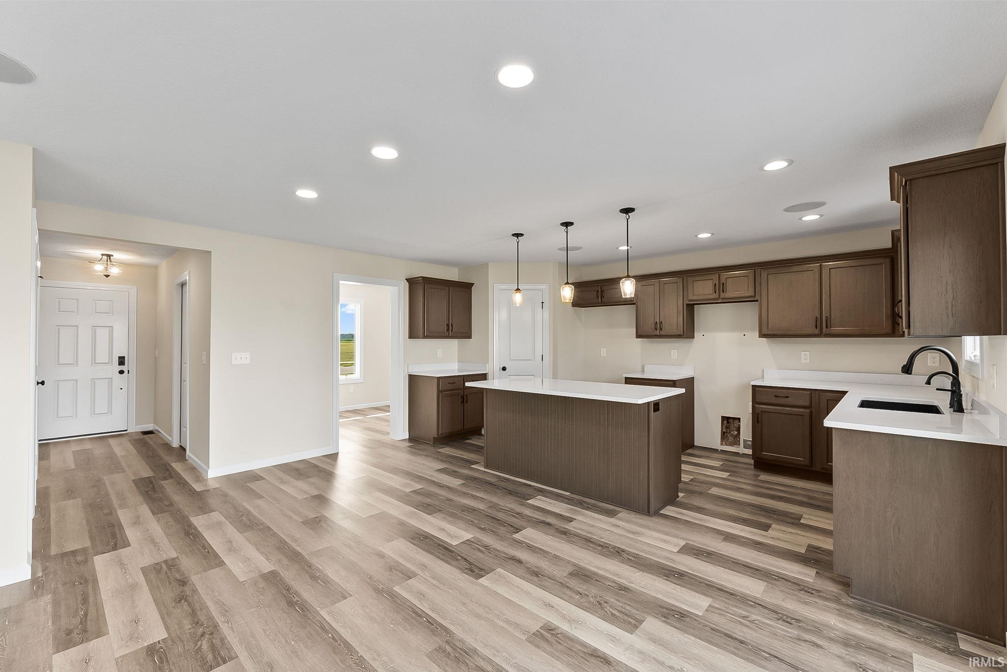 Open-concept kitchen featuring wood-finish flooring, recessed lighting, a central island with pendant lights, dark wood cabinetry, and white countertops