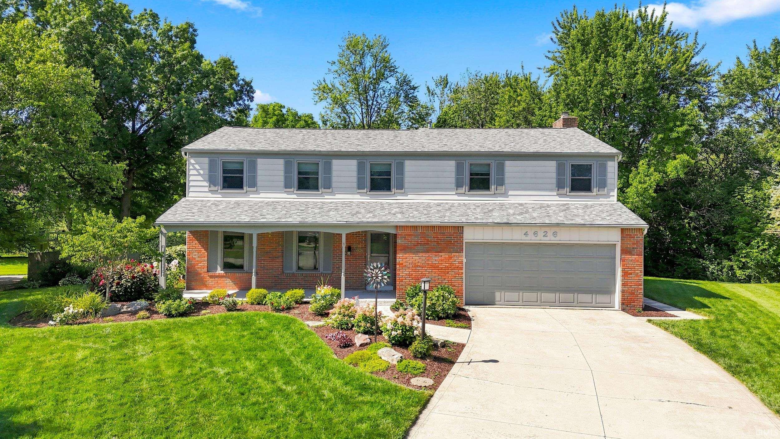 Brick Colonial two-story home featuring a clean driveway approach, attached two-car garage with paneled doors, and a manicured front lawn bordered by mature landscaping